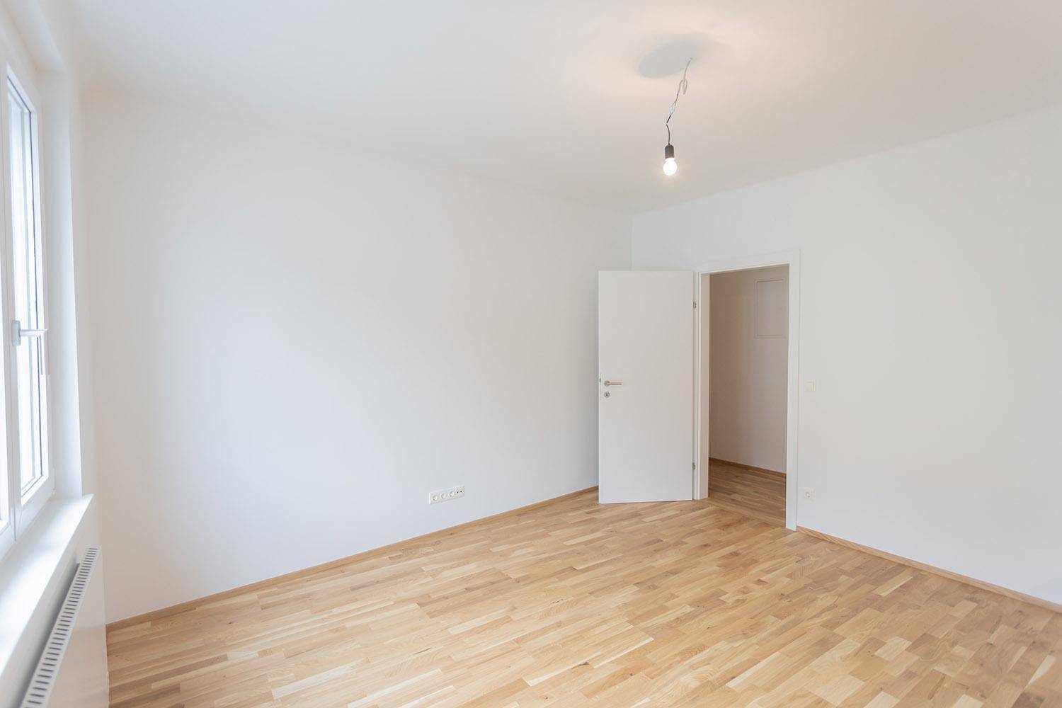 Leeres Zimmer mit hellem Holzboden und großem Fenster, das viel Tageslicht hereinlässt.