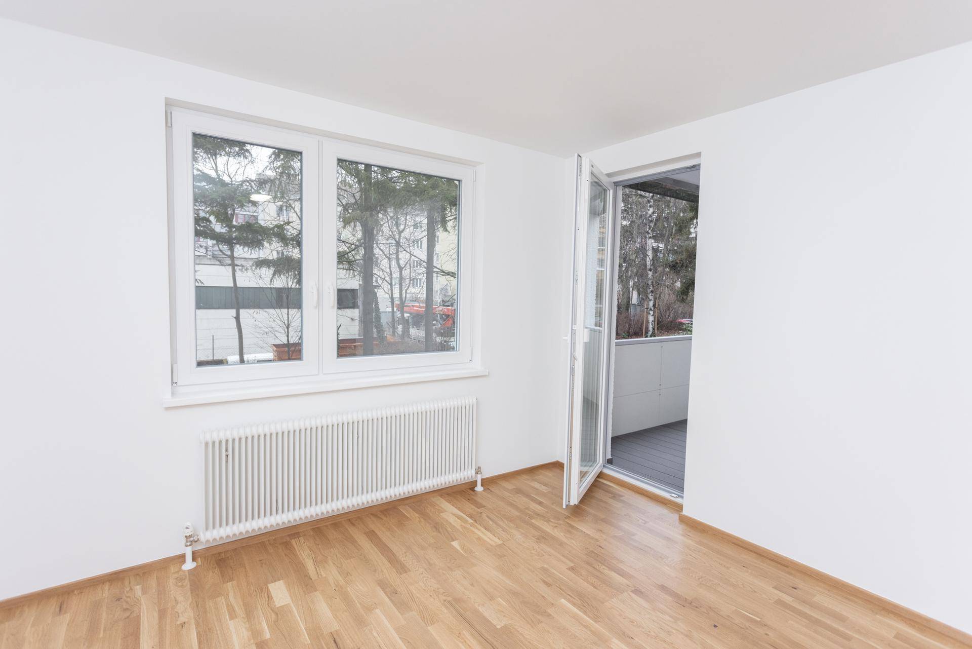 Heller Raum mit Holzboden, großem Fenster und Zugang zu einem Balkon.