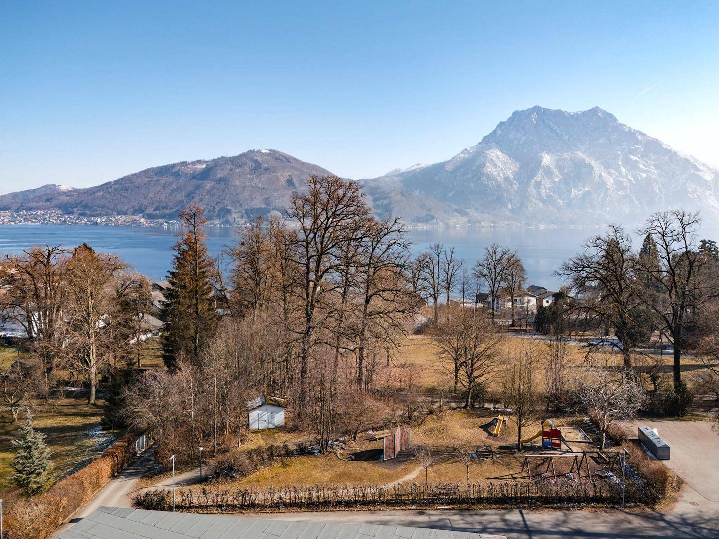 Panoramablick auf den See, majestätische Berge und einen Spielplatz mit kahlen Bäumen.