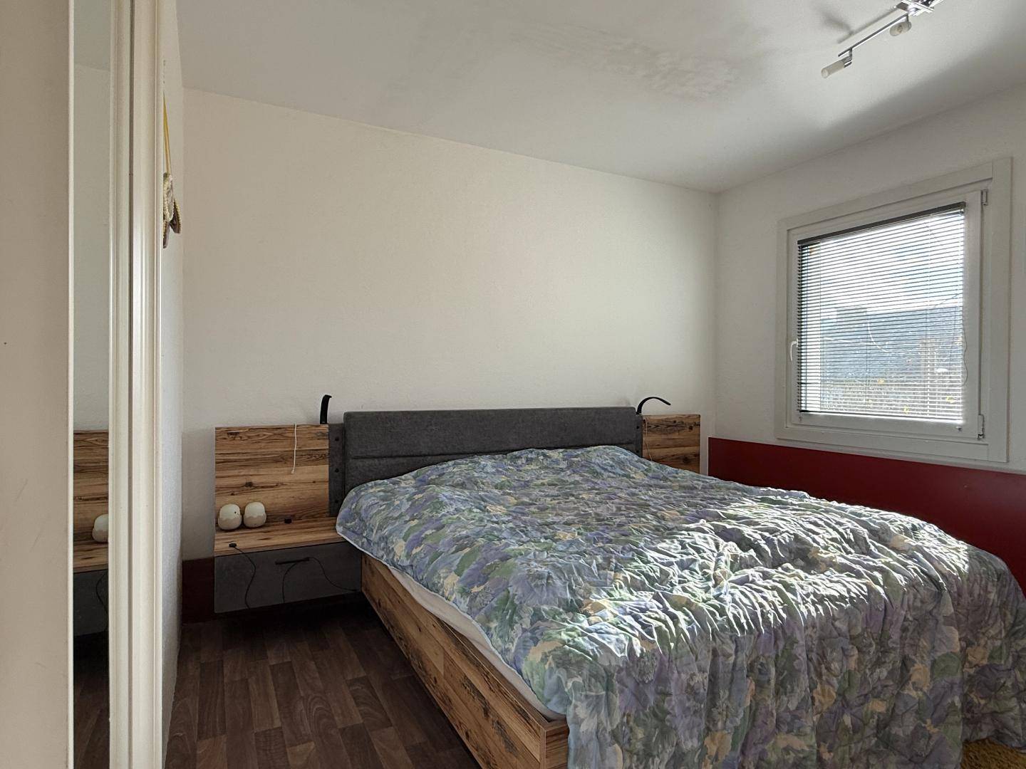 Schlafzimmer mit modernem Holzbett und Fenster mit Jalousien für Privatsphäre.