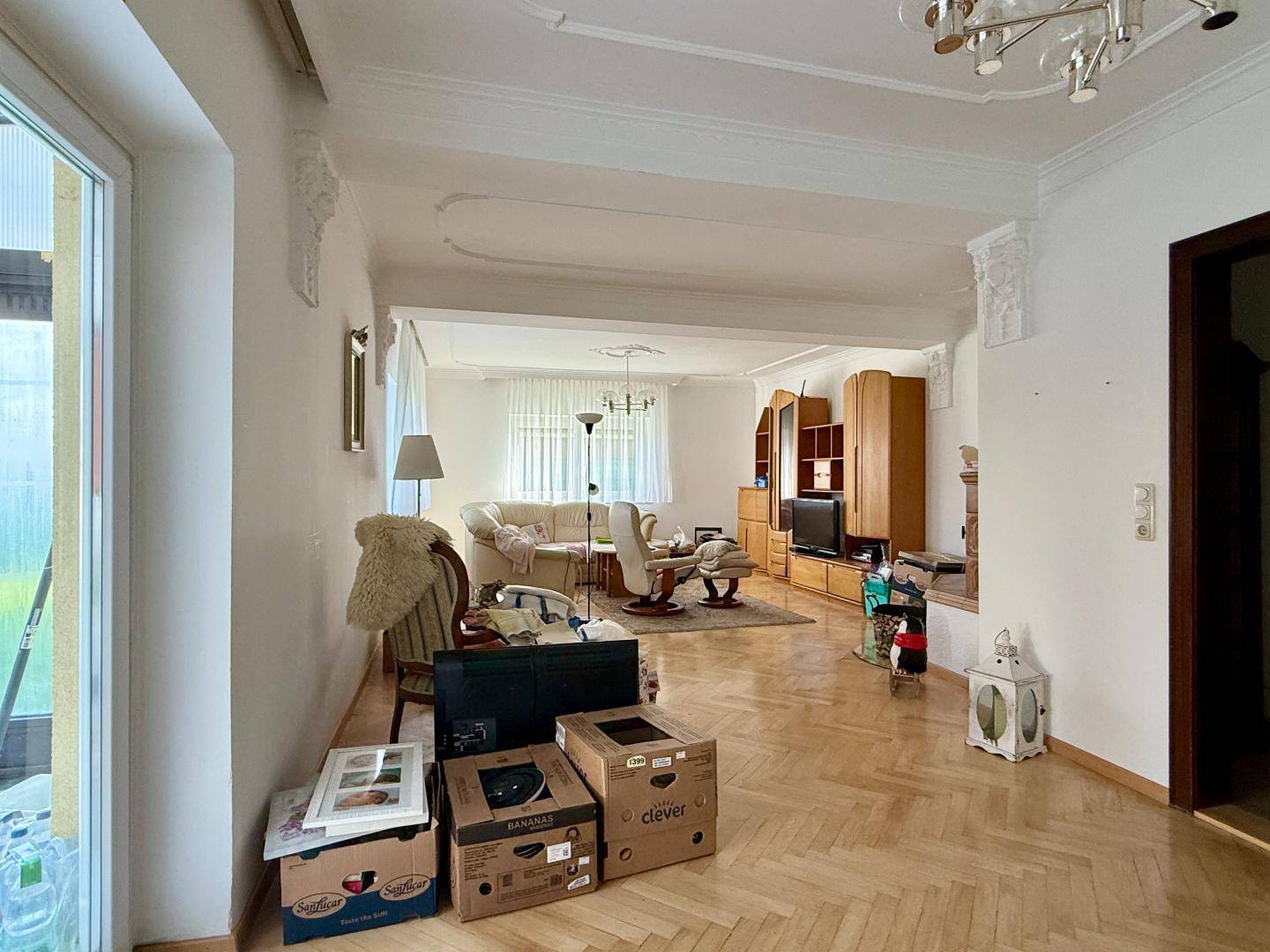 Geräumiges Wohnzimmer mit Parkettboden, Stuckelementen und Blick in den Essbereich.