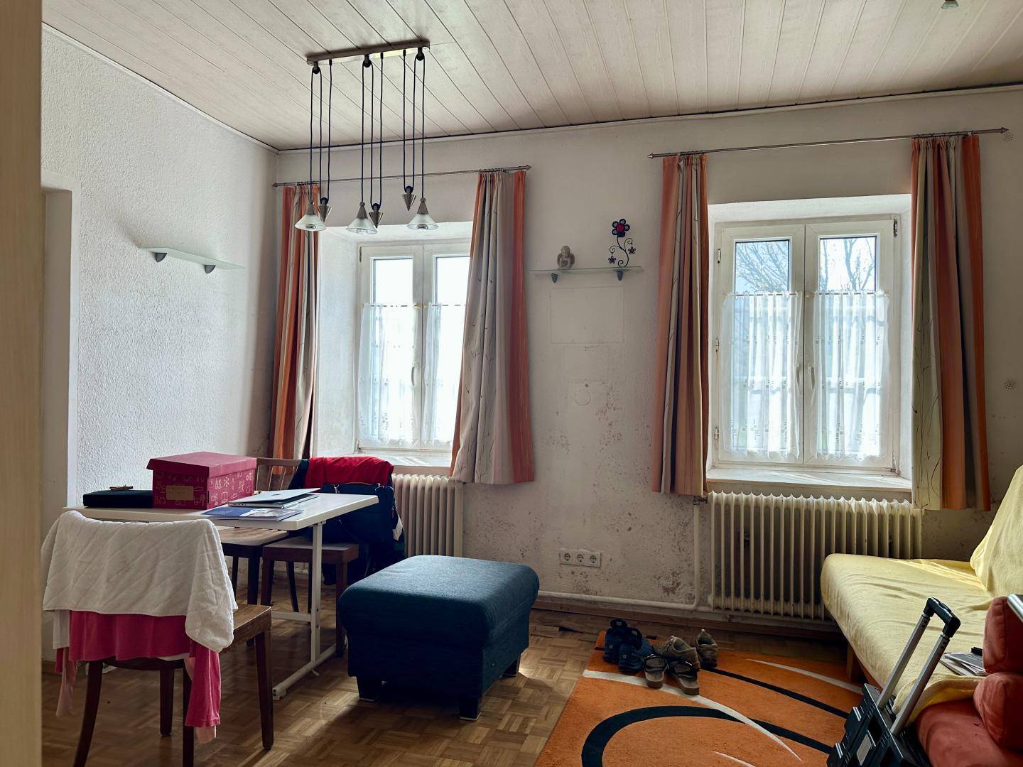 Wohnraum mit Holzbalkendecke, Parkettboden und zwei Fenstern, die viel Licht hereinlassen.