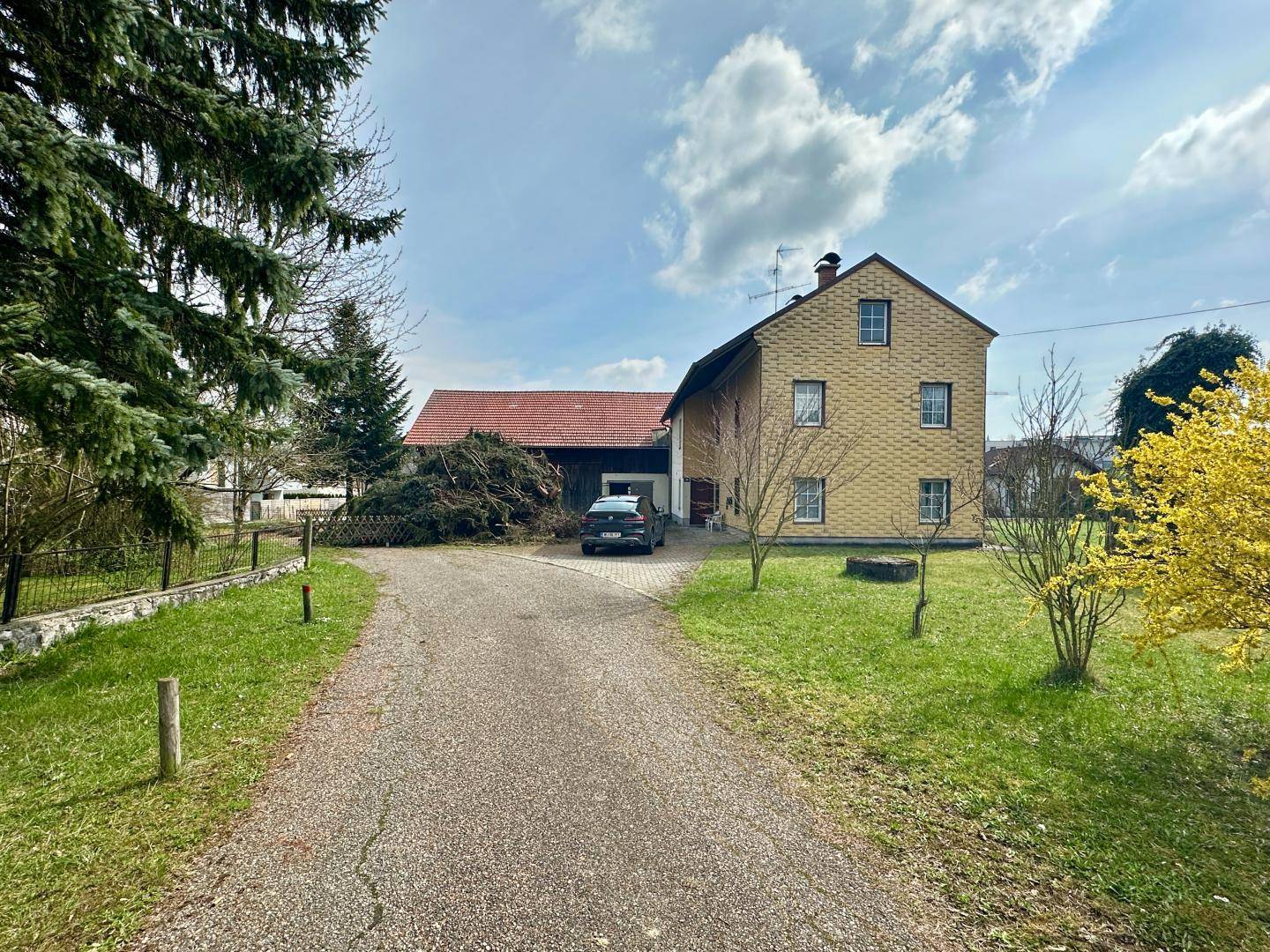 Kiesauffahrt vor einem traditionellen Haus mit Scheune und blühenden Sträuchern.