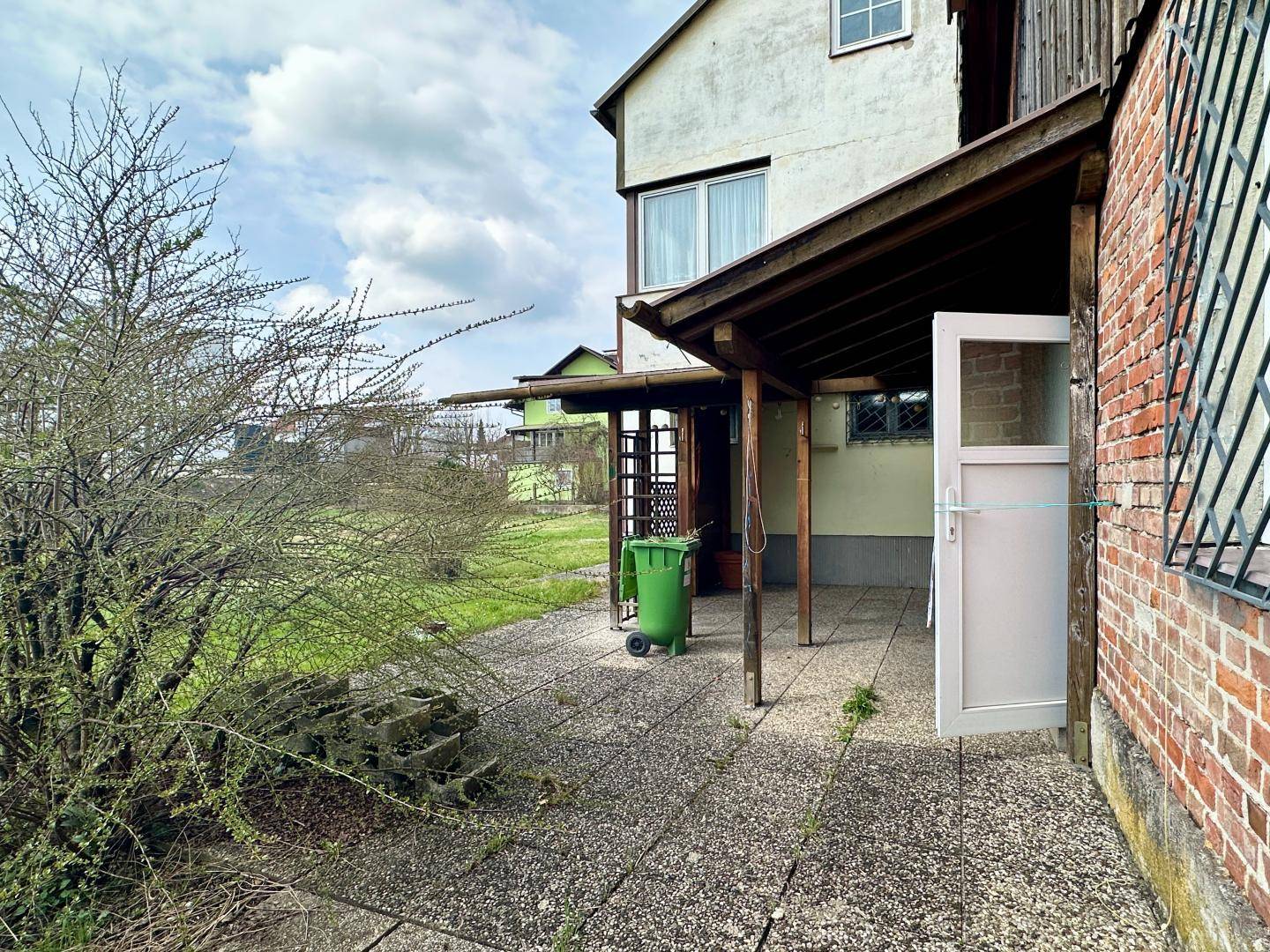 Gepflasterte Terrasse neben dem Haus mit Grünfläche und Mülltonne.