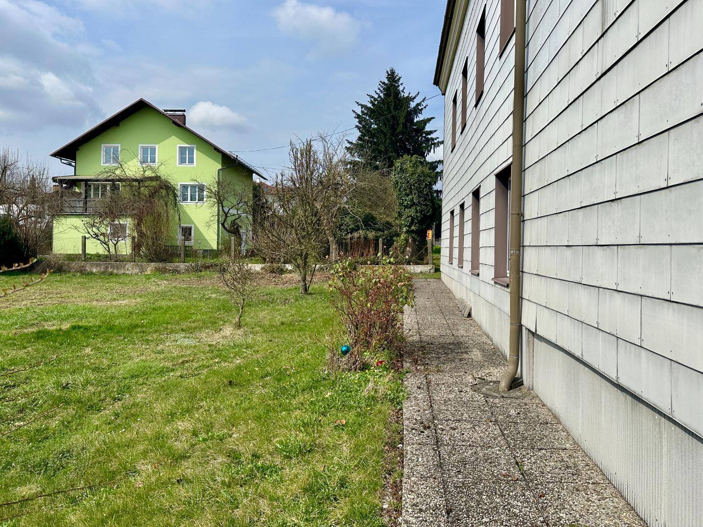 Seitenansicht eines Hauses mit grünem Nachbargebäude und gepflegter Rasenfläche.