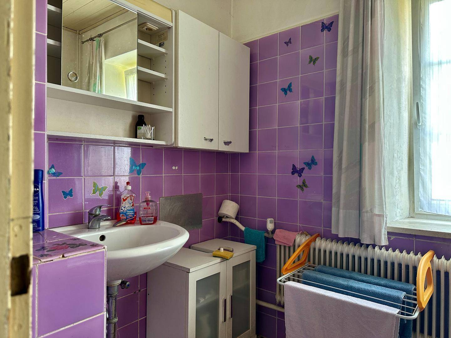 Badezimmer mit lila Fliesen, Waschbecken und Fenster, das einen 80er-Jahre-Stil aufweist.
