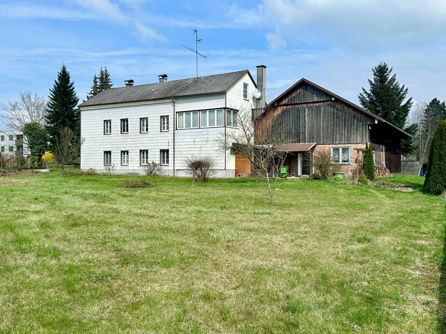 Ansicht eines Hauses mit Scheune und großem Grundstück unter einem bewölkten Himmel.