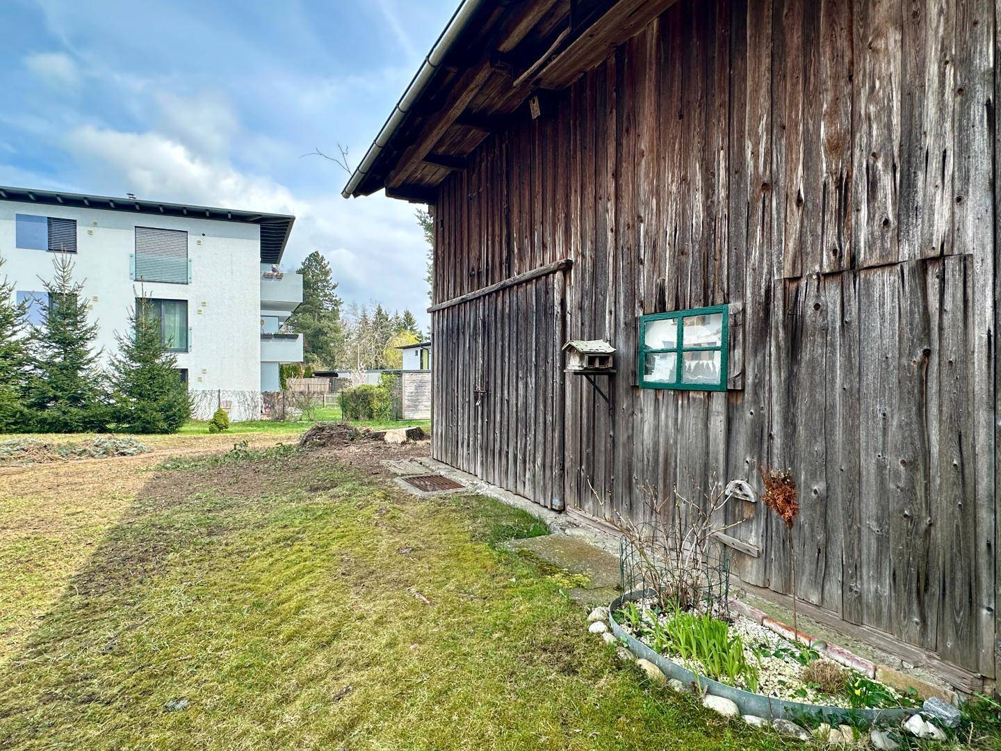 Rustikale Holzscheune mit kleinem Fenster und Blumenbeet im Vordergrund.