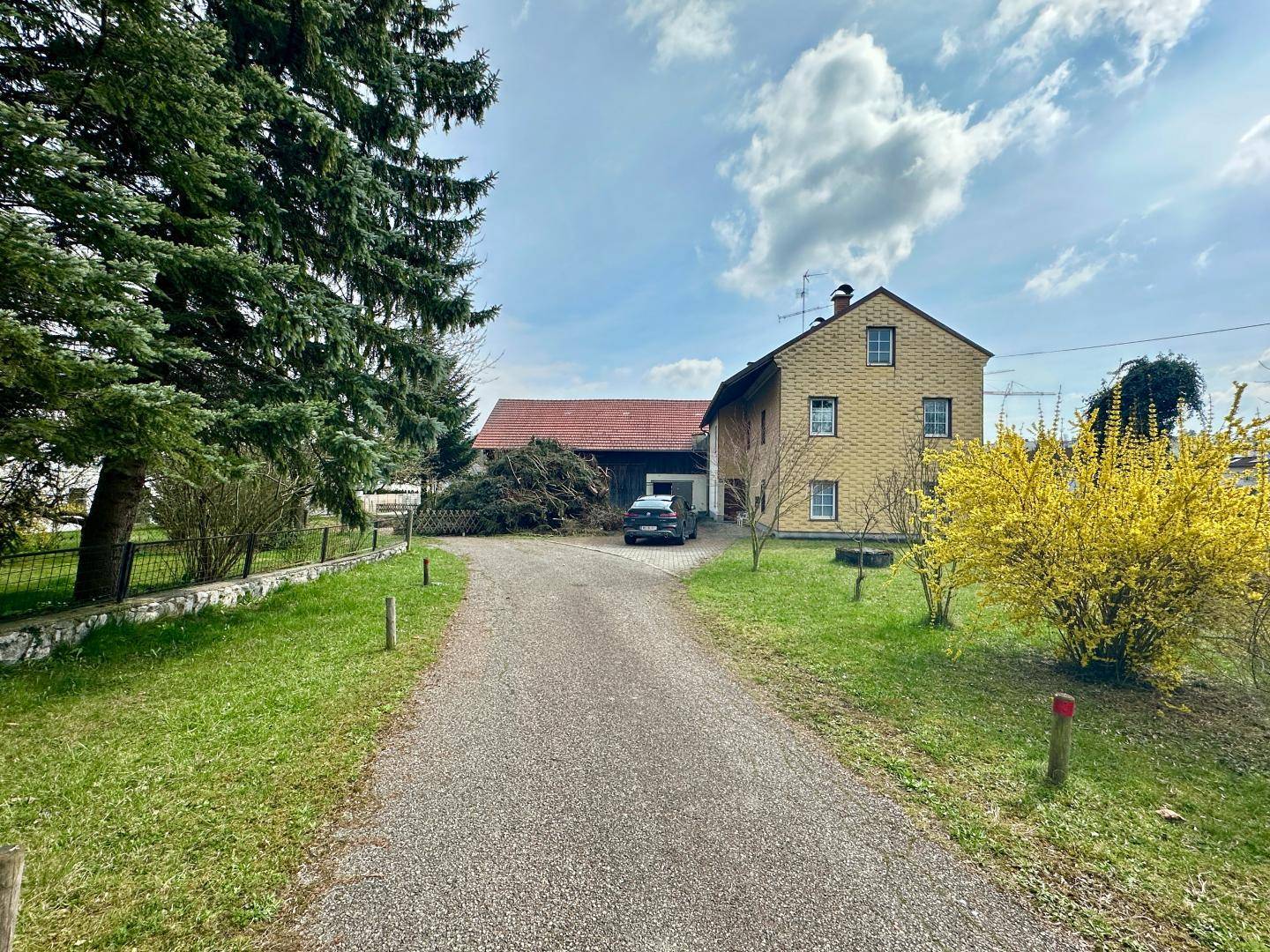 Kiesauffahrt führt zu einem traditionellen Haus mit angrenzender Scheune und hohen Bäumen.