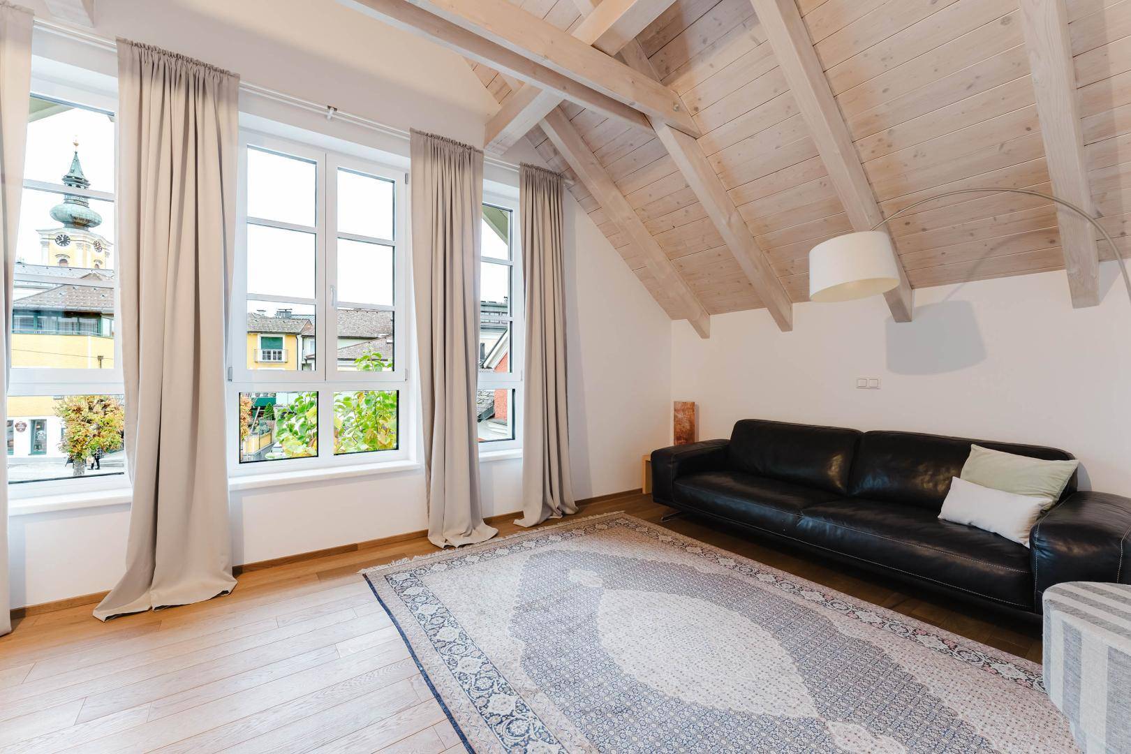 Geräumiges Wohnzimmer im Dachgeschoss mit Holzbalken, Teppich und Stadtblick.