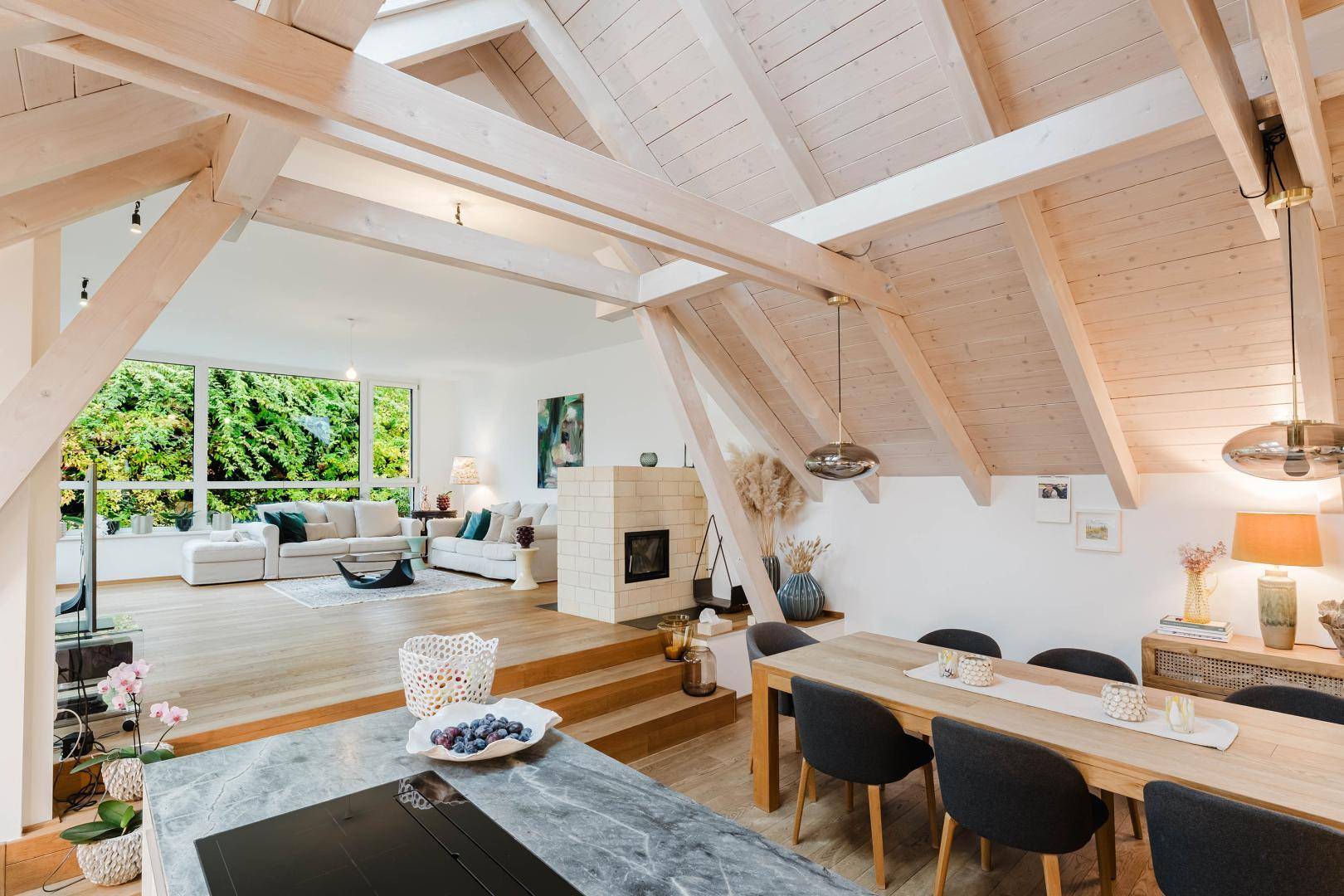 Offener Wohn- und Essbereich mit Holzbalken, Kamin und großen Fenstern zum Grünen.