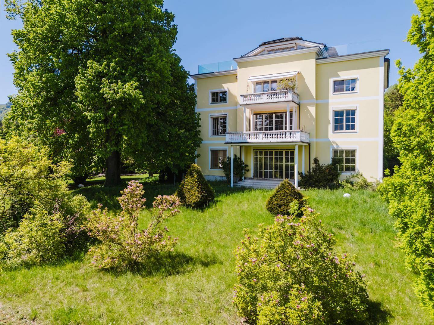 Weitläufige Luftaufnahme der gelben Villa mit ihrem großen, grünen Garten und altem Baumbestand.