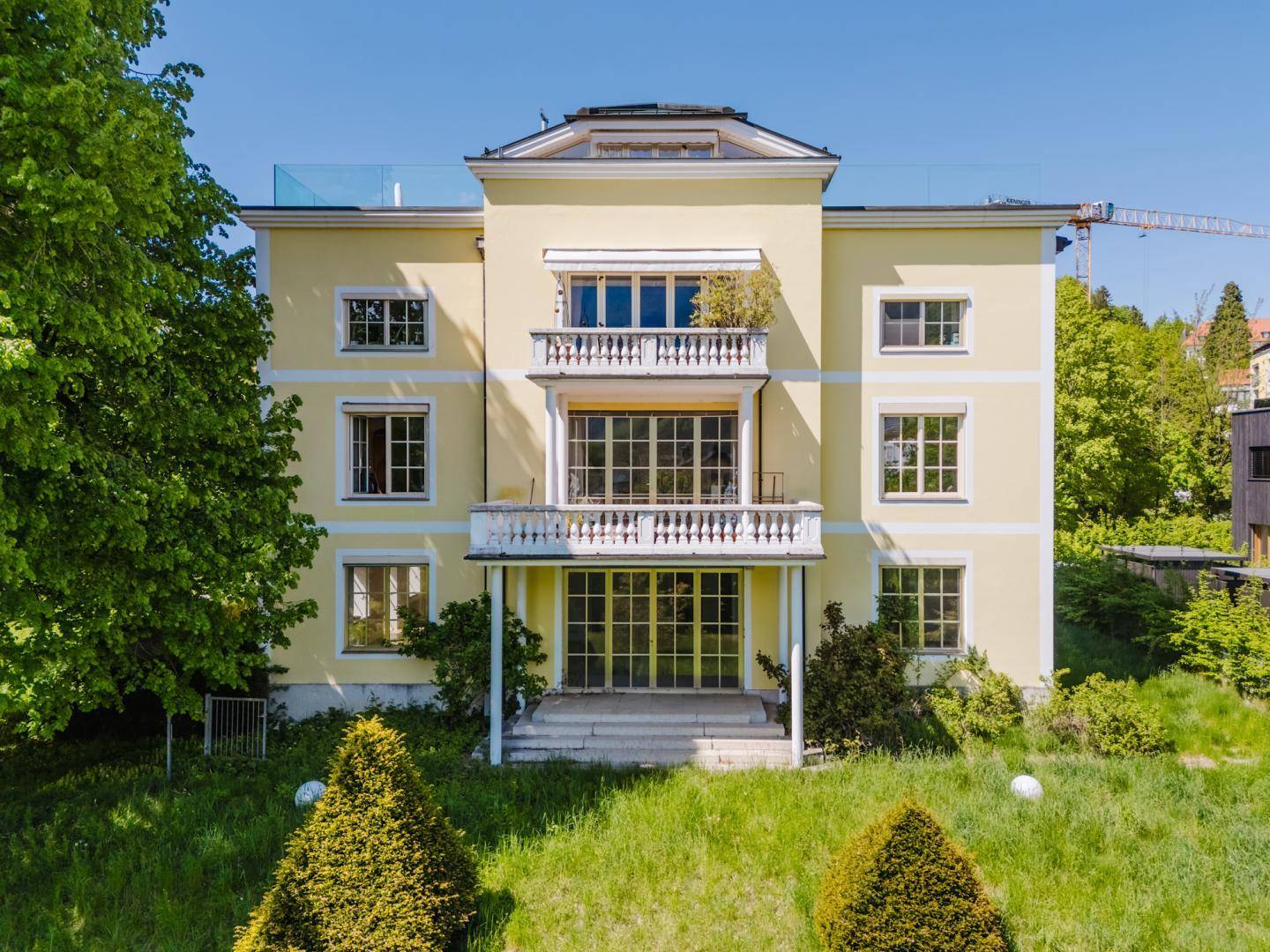 Luftaufnahme der gelben Villa von vorne, umgeben von einem weitläufigen Garten und Bäumen.