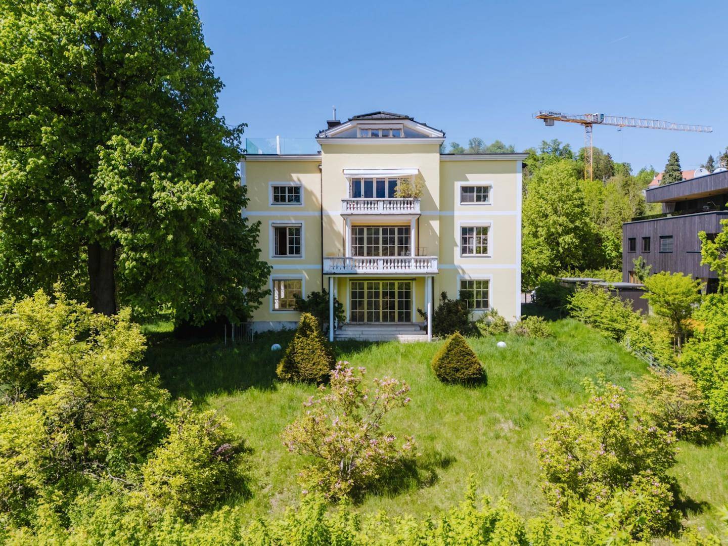 Luftaufnahme der gelben Villa von vorne, umgeben von einem weitläufigen Garten und Bäumen.