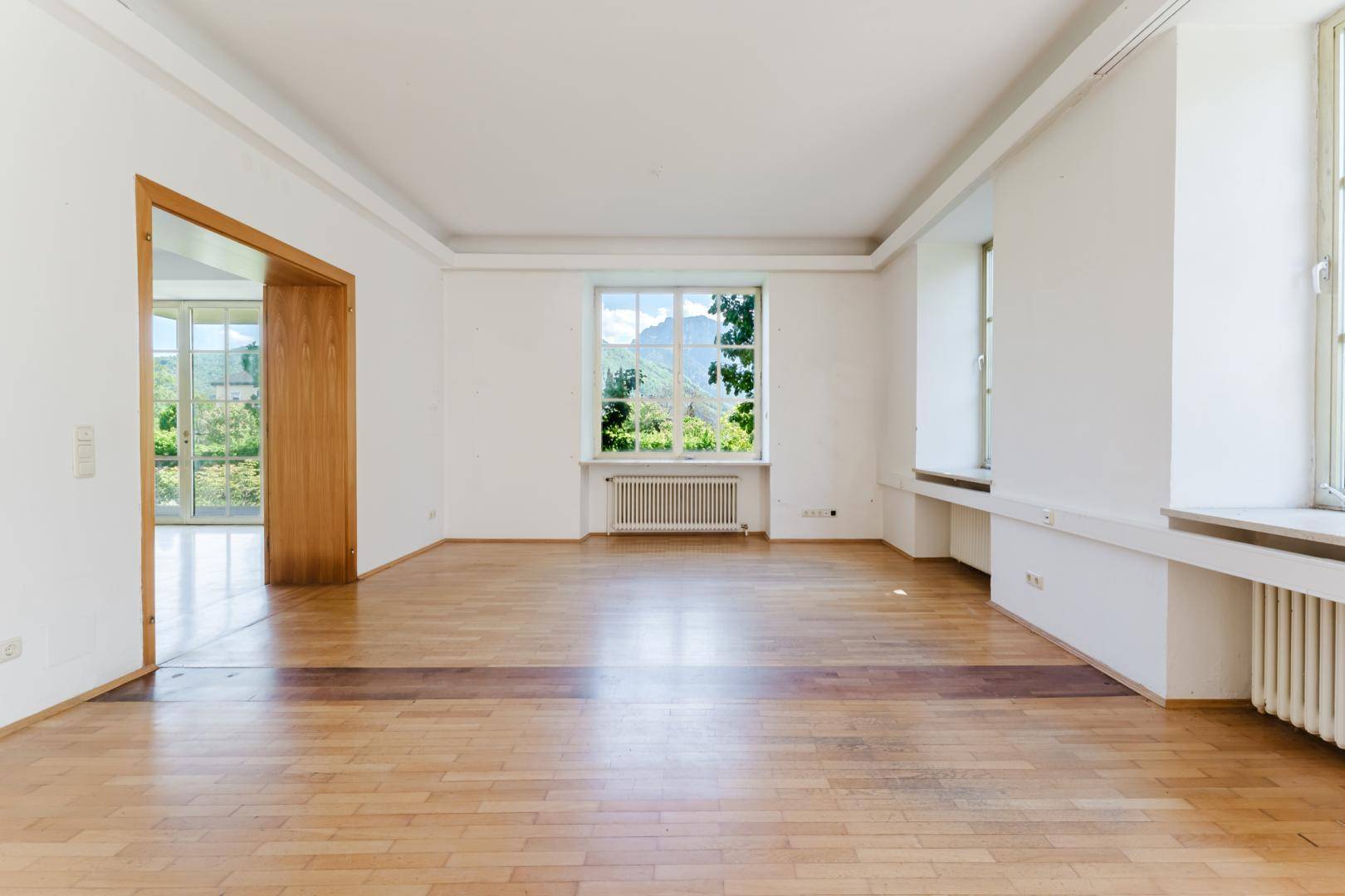 Heller Raum mit Fischgrätparkettboden, einem Fenster mit Bergblick und Zugang zu weiteren Zimmern.