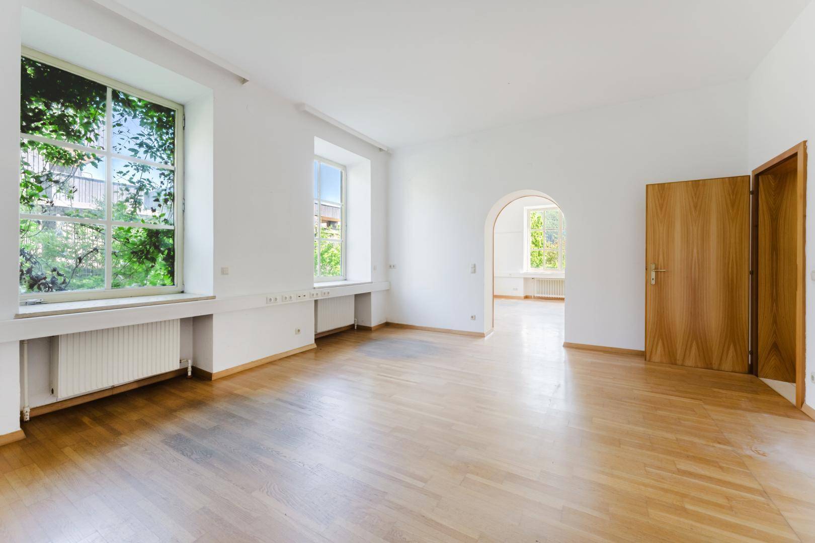 Geräumiger Raum mit Parkettboden, großen Fenstern und einem eleganten Rundbogen zum nächsten Bereich.
