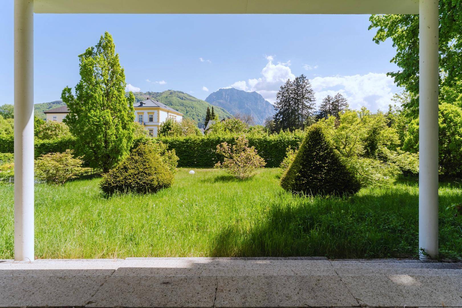 Blick von einer überdachten Loggia auf einen weitläufigen grünen Garten und die majestätischen Berge.