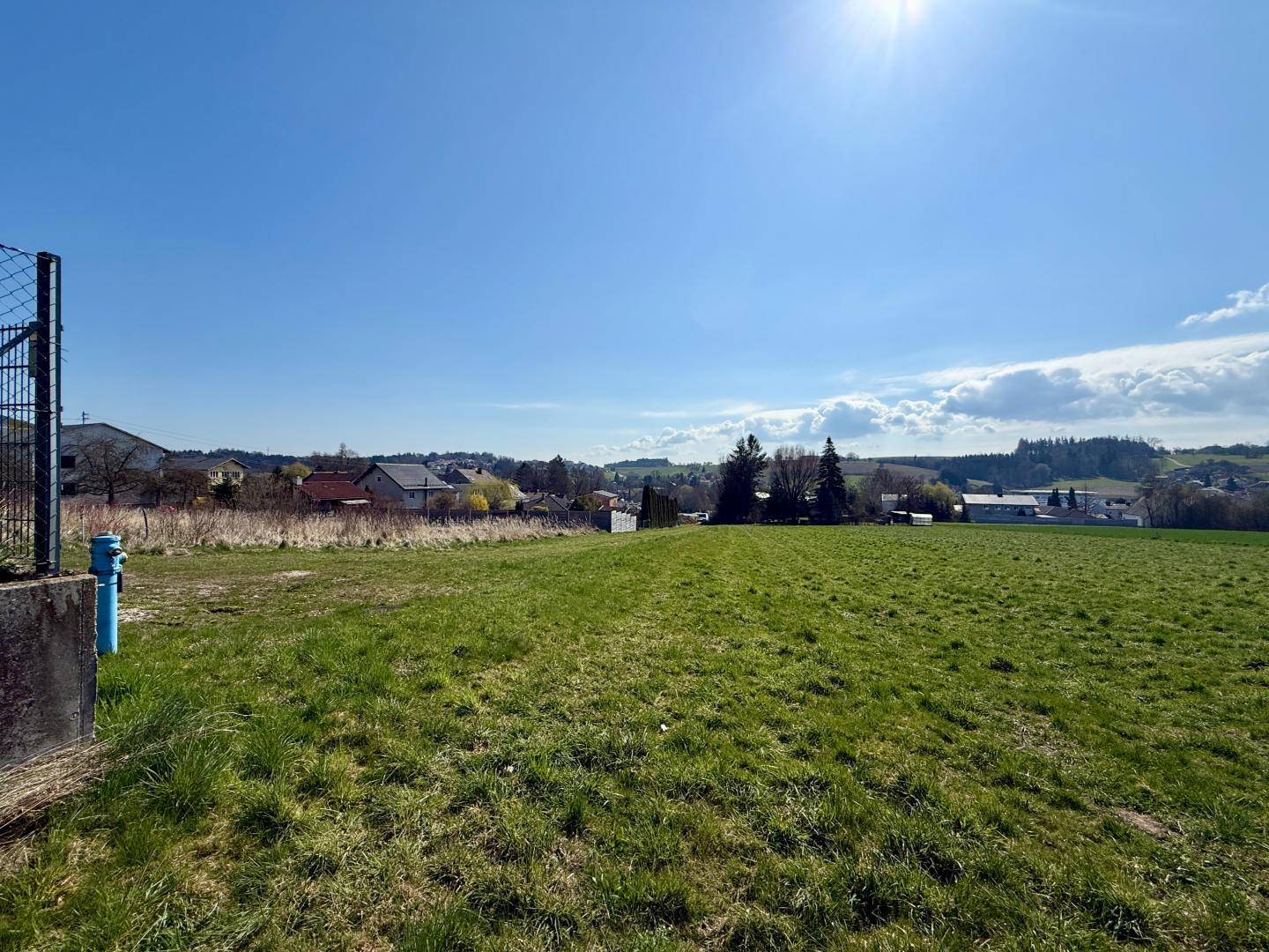 Grünes Feld mit einem Zaun und Hydranten im Vordergrund, dahinter eine ländliche Siedlung.