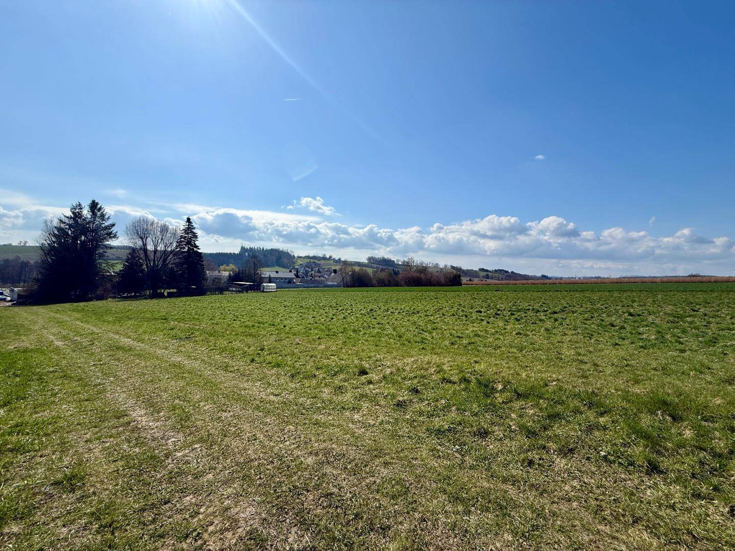 Sonnige, weite Grünfläche mit Blick auf eine entfernte Siedlung und bewaldete Hügel.