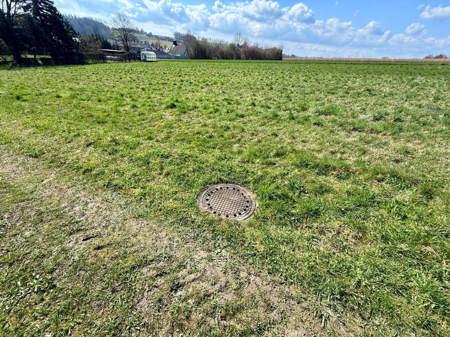Grüne Wiese mit einem Kanaldeckel im Vordergrund und einem weiten Blick in die ländliche Umgebung.