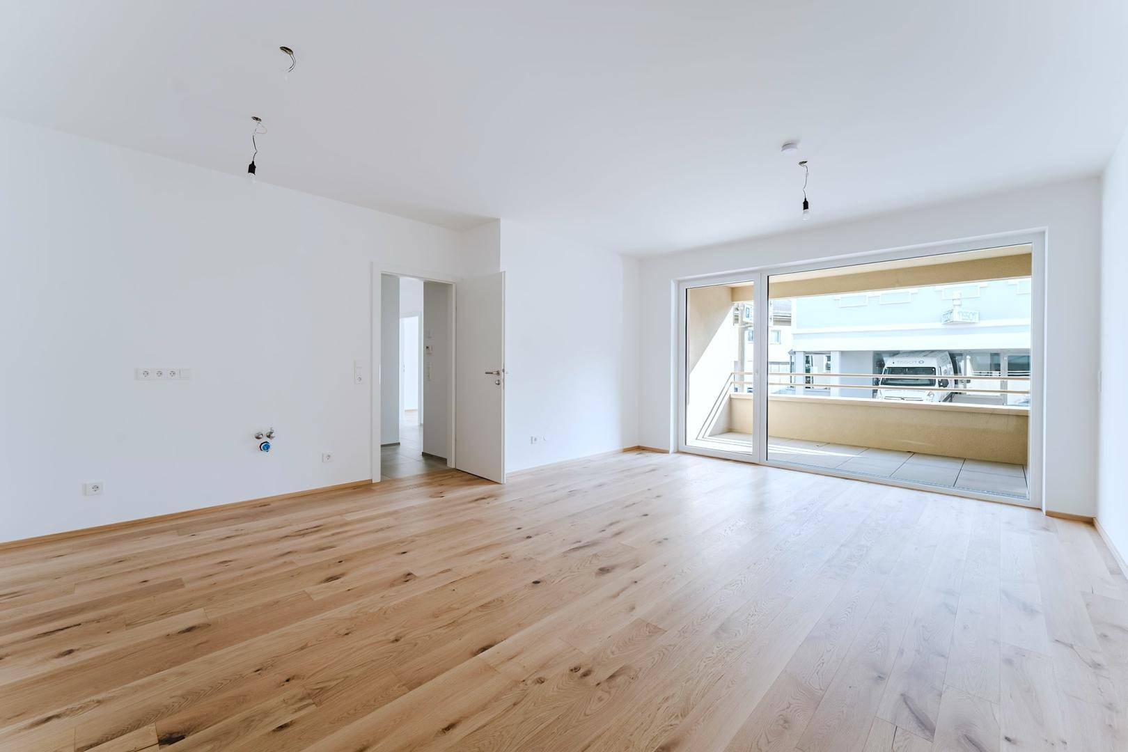 Heller, leerer Raum mit Holzboden, Balkonzugang und Blick auf den Flur.