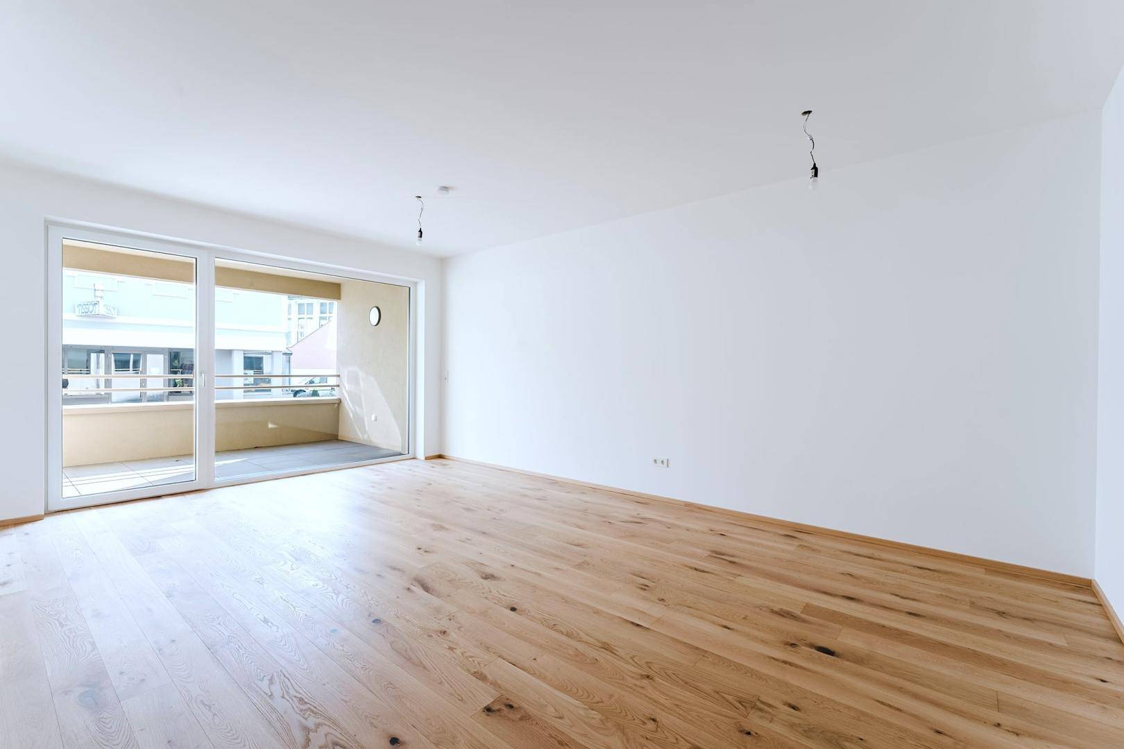 Geräumiges, leeres Wohnzimmer mit hellem Holzboden und großem Balkonzugang.