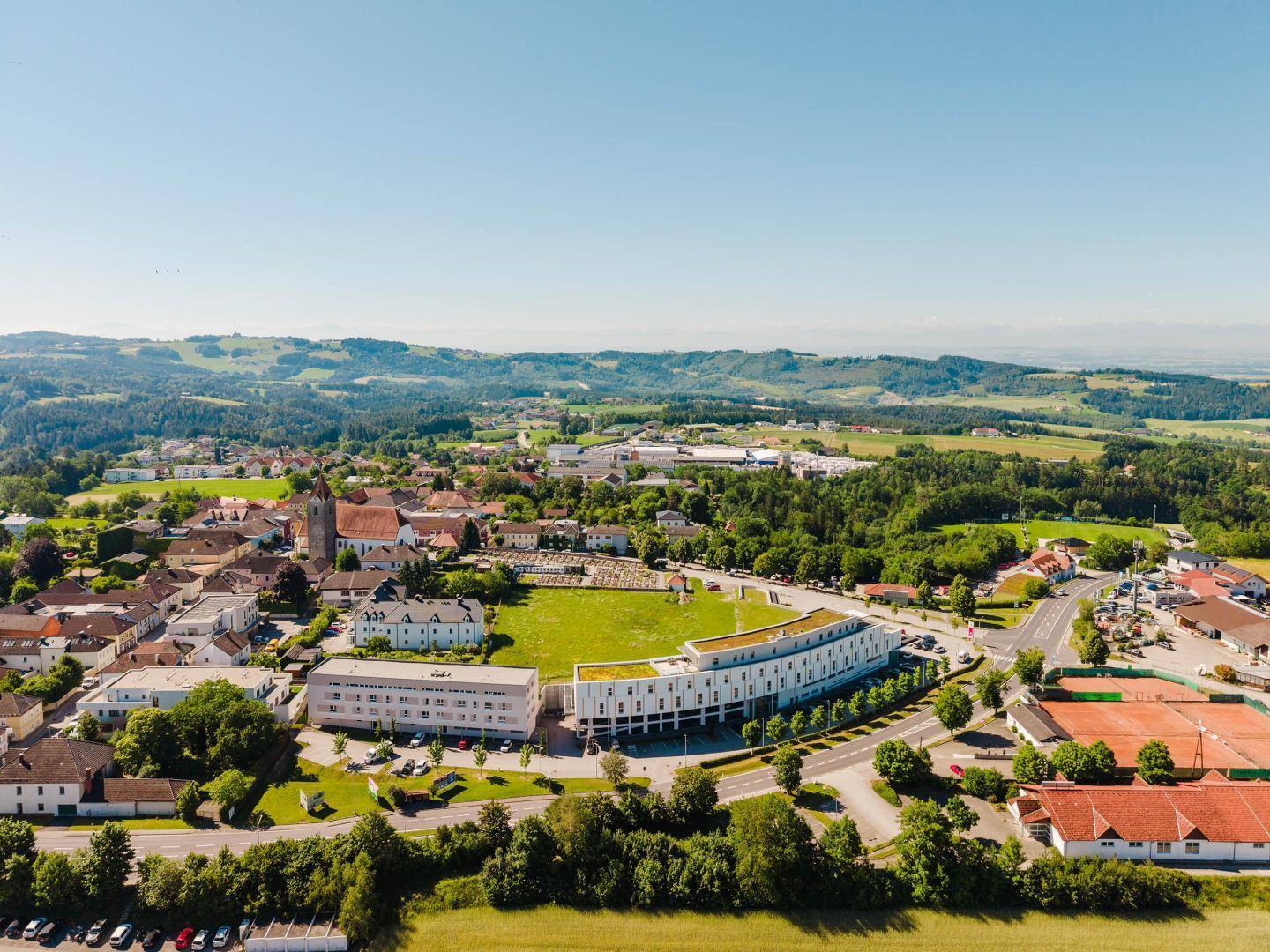 Weitläufige Luftaufnahme der Stadt mit modernem Gebäude, Kirche und Tennisplätzen.