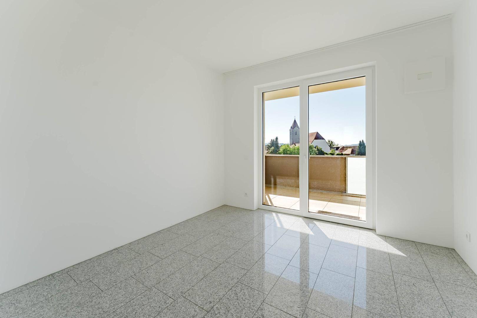 Heller Raum mit Fliesenboden und Zugang zum Balkon mit Blick auf die Kirche.