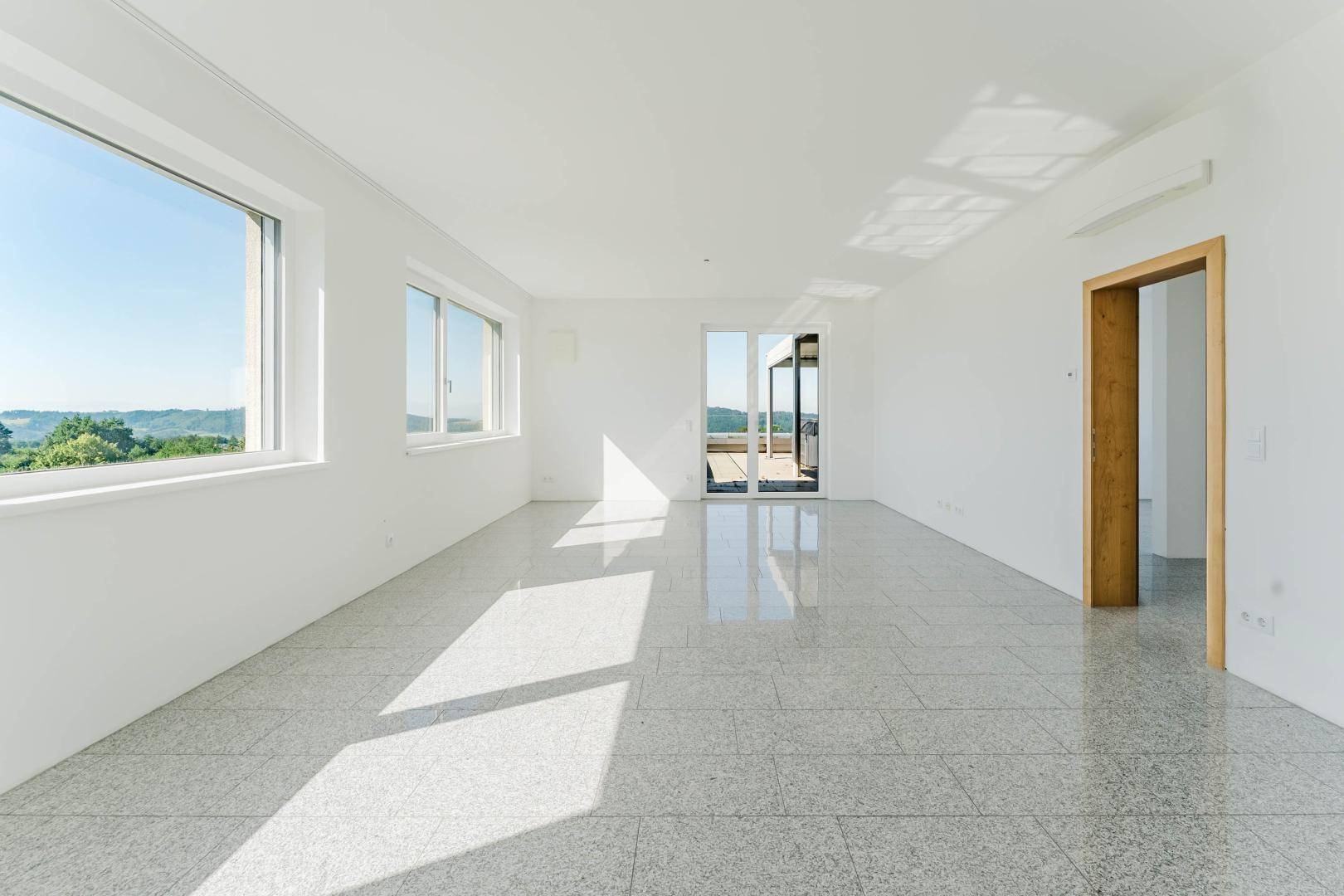 Helles, leeres Zimmer mit großen Fenstern, die einen weiten Blick ins Grüne bieten und Zugang zur Terrasse.