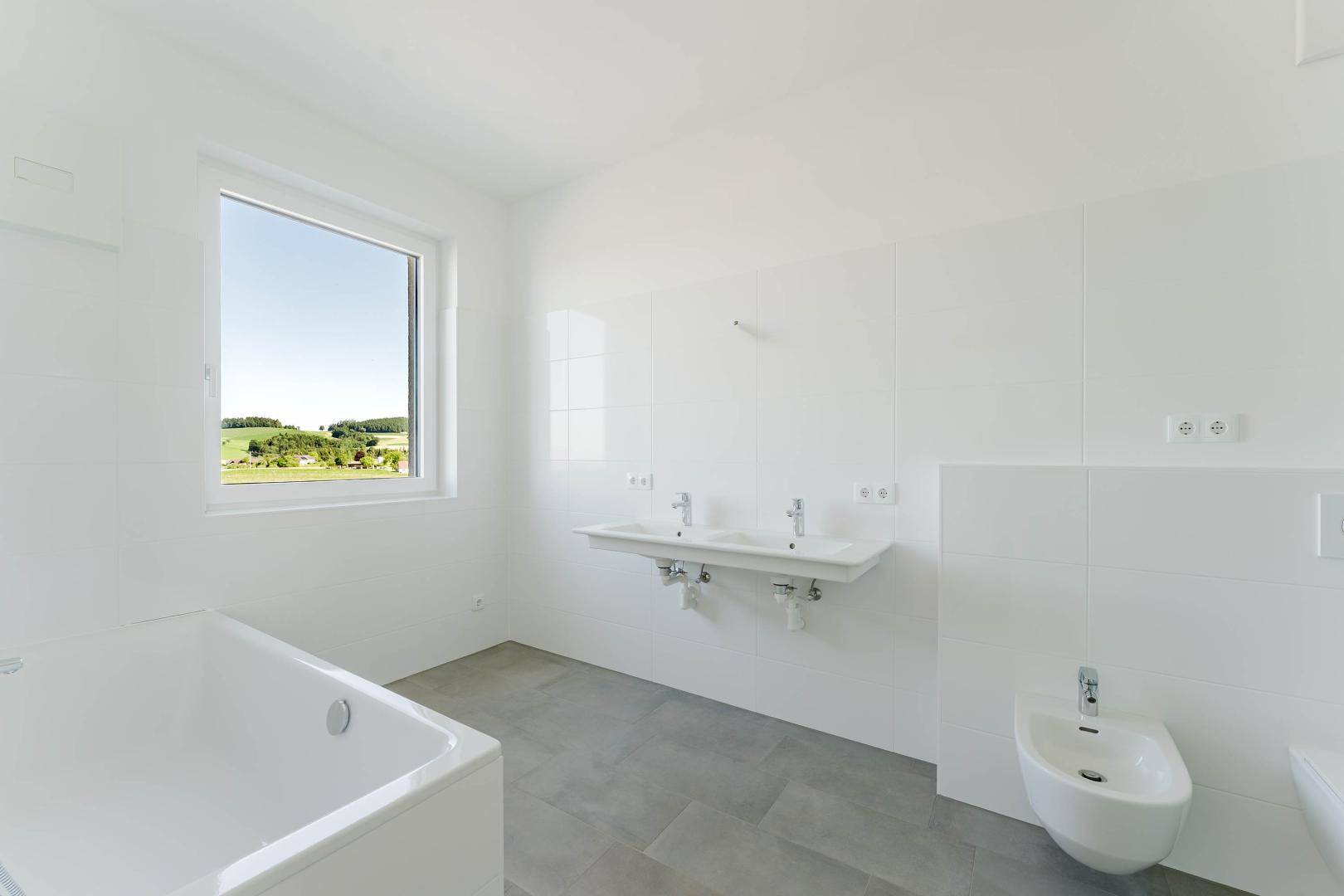 Modernes Badezimmer mit weißen Fliesen, Doppelwaschbecken, Badewanne und Bidet, mit Fensterblick ins Grüne.