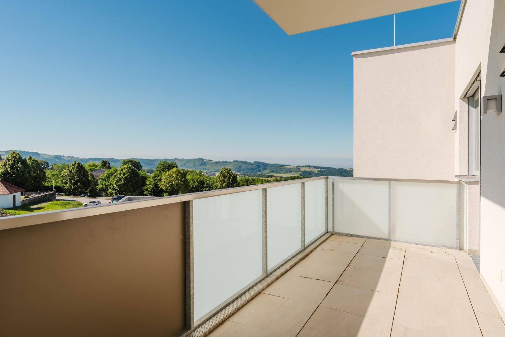 Heller Balkon mit Glasgeländer und weitem Blick über die grüne Umgebung.