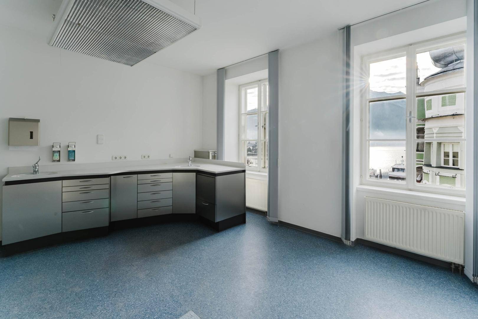 Professioneller Büroraum mit blauer Bodenfläche, Edelstahlmöbeln und hellem Fenster.