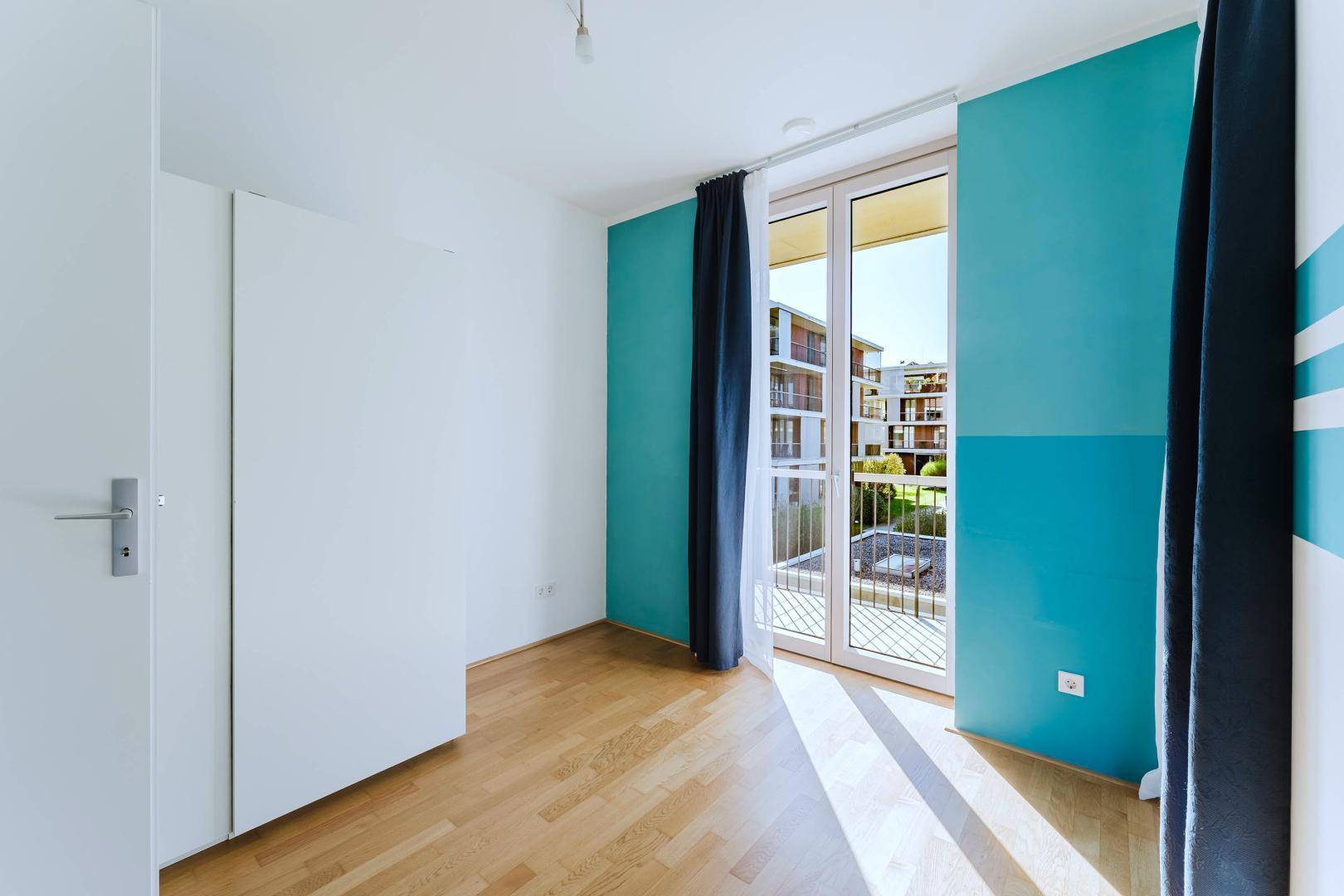 Helles Zimmer mit Holzboden, blauer Akzentwand und Zugang zu einem Balkon mit Blick auf andere Gebäude.