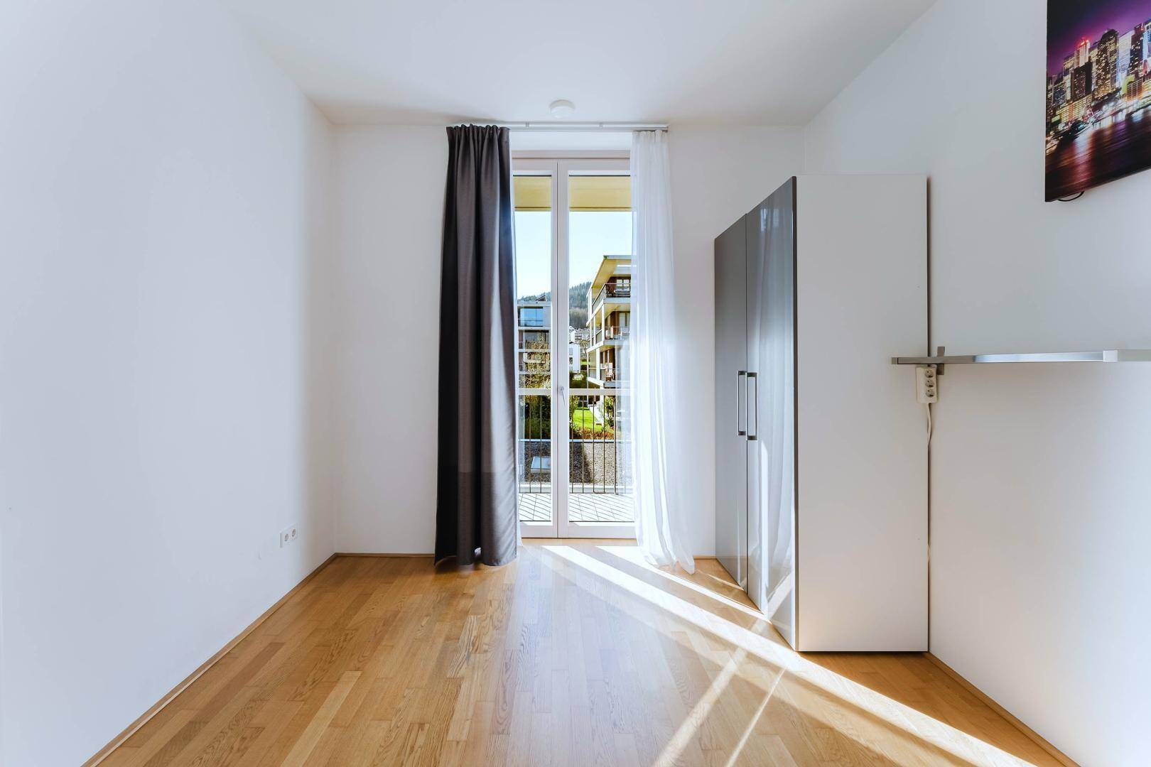 Helles Schlafzimmer mit Holzboden, weiß gestrichenen Wänden und Balkonzugang.