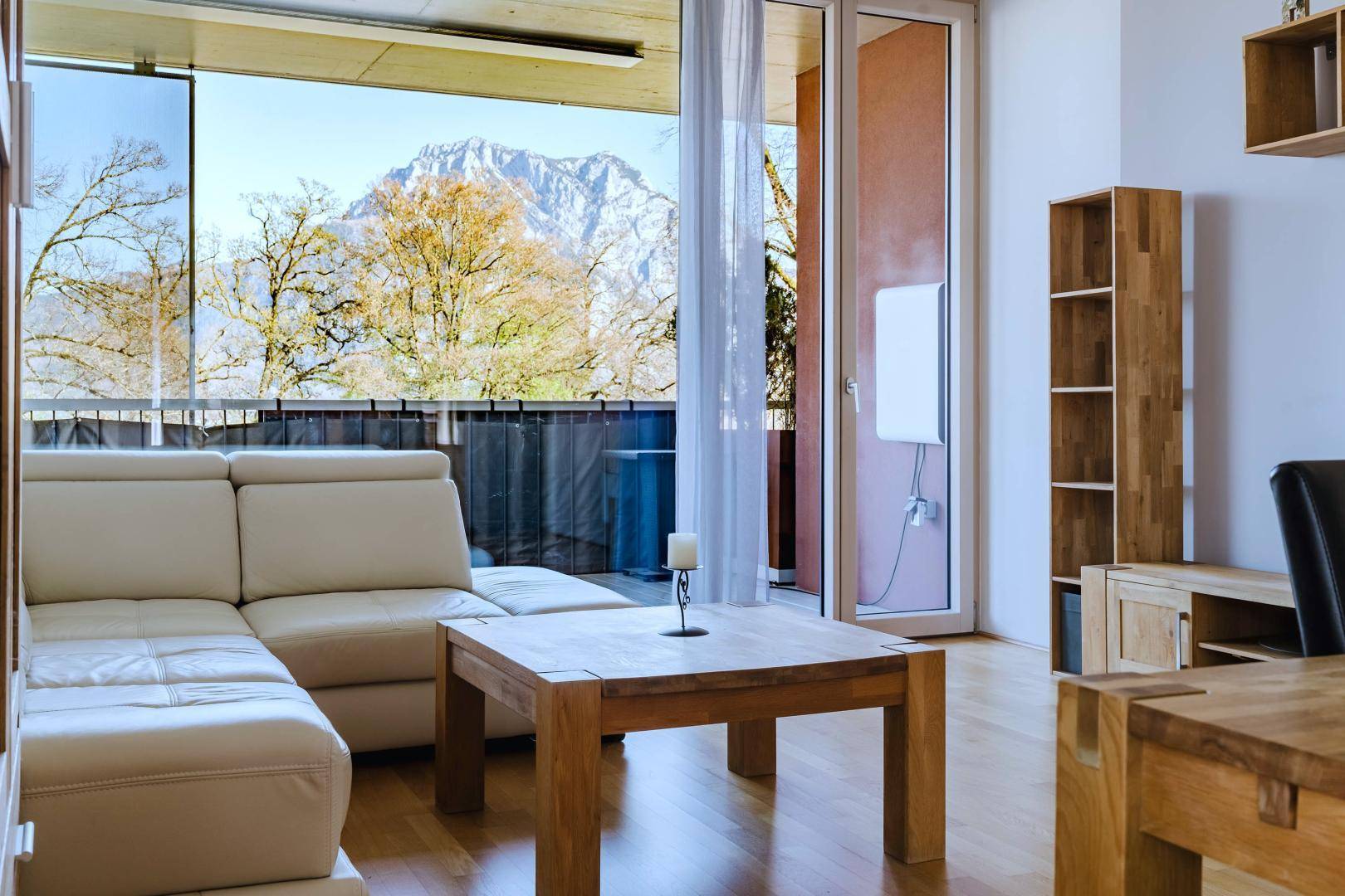 Lichtdurchflutetes Wohnzimmer mit beiger Ledercouch, Holztisch und weitem Bergblick vom Balkon.