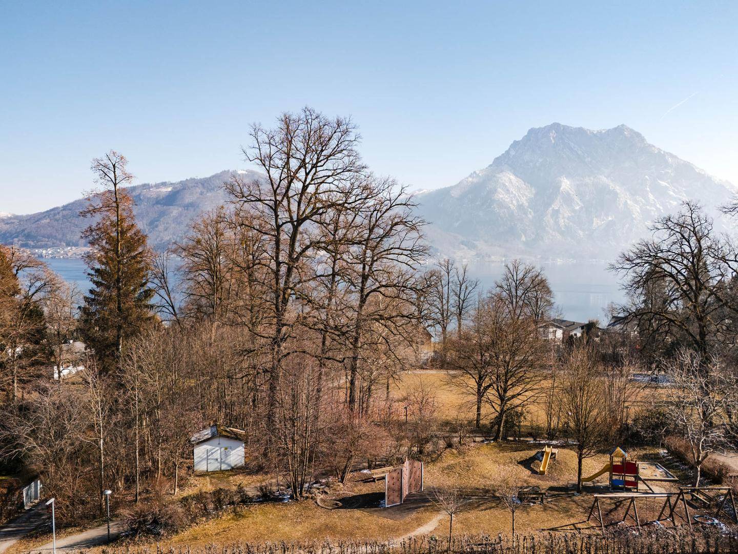 Weitläufiger Blick auf einen Spielplatz, Bäume, See und Berge.