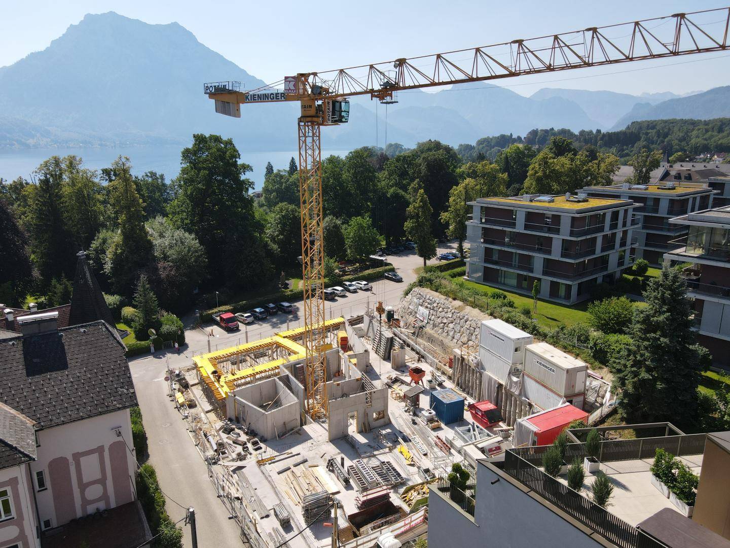Luftaufnahme einer Baustelle mit Kran und Rohbauarbeiten, umgeben von See und Bergen.
