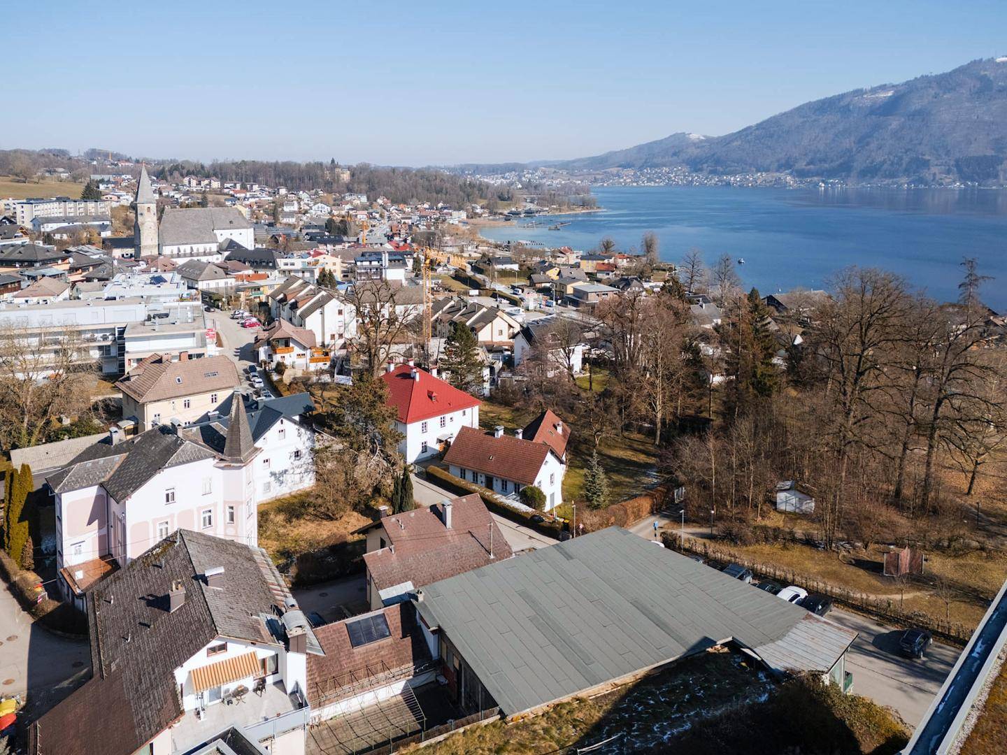 Luftaufnahme einer Stadt mit Wohngebäuden, Kirche, See und Bergen in der Ferne im Winter.