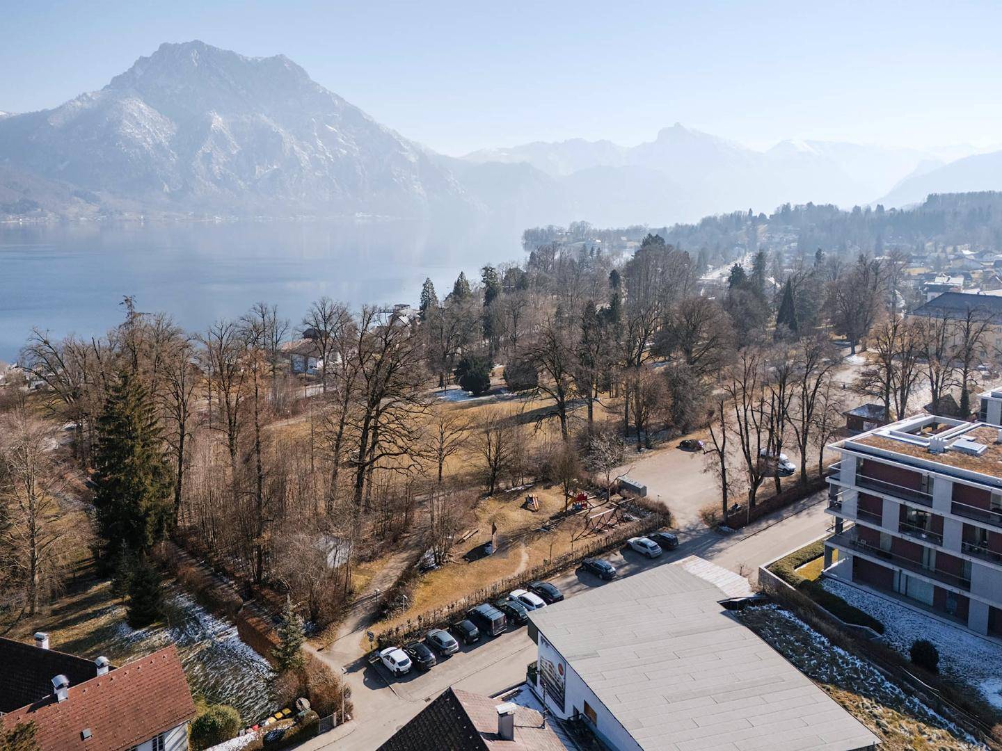 Luftaufnahme eines Parks mit Spielplatz, See und schneebedeckten Bergen im Hintergrund an einem sonnigen Tag.