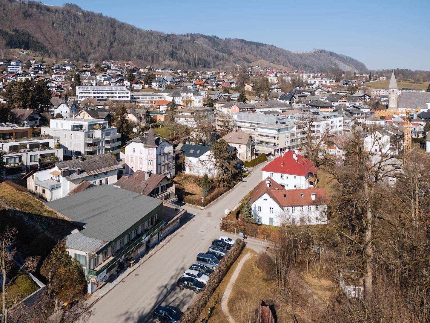 Luftaufnahme einer Stadt mit Wohngebäuden, Kirche und Bergen in der Ferne unter blauem Himmel.
