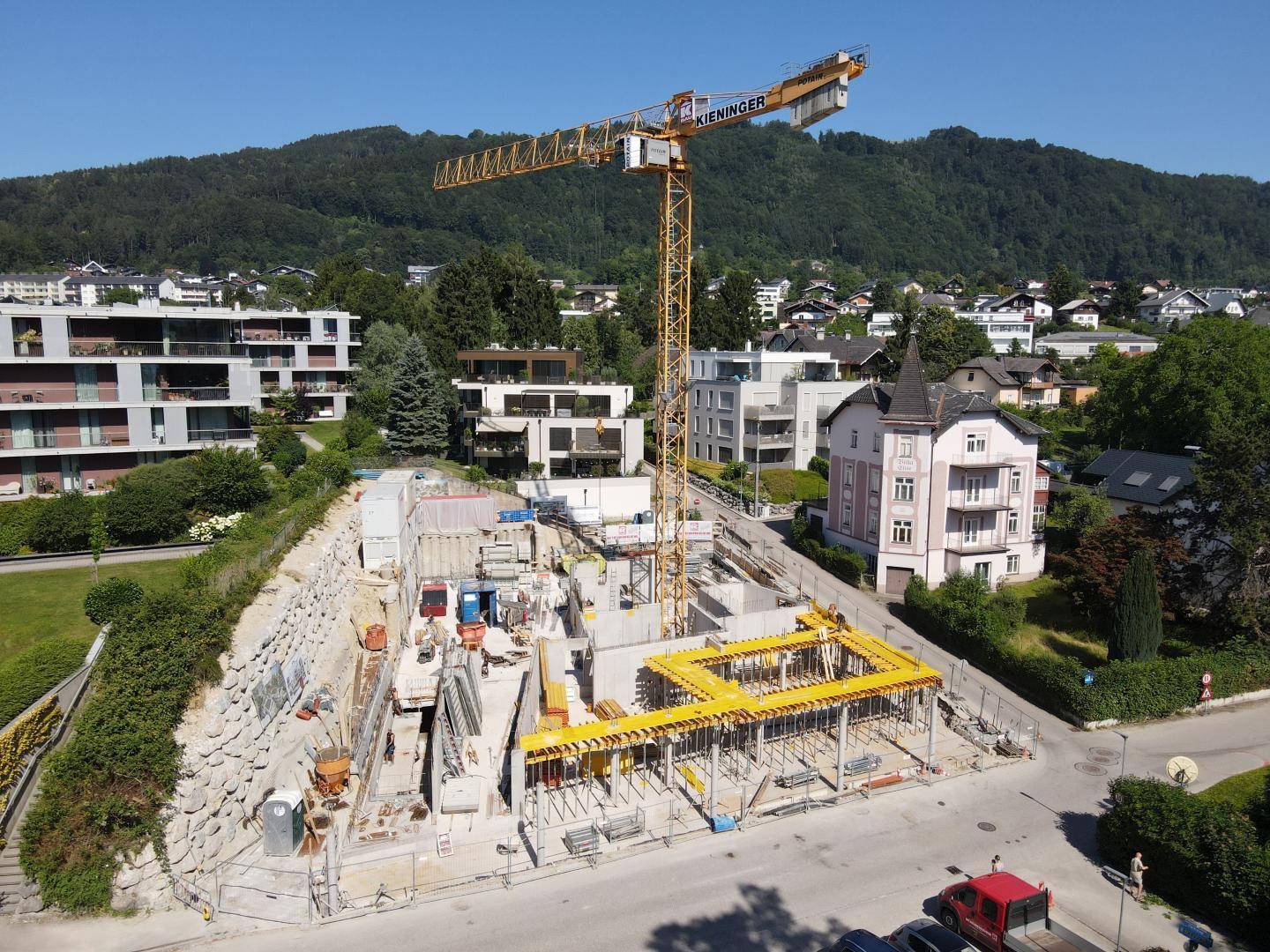 Baustelle mit Kran und umliegenden Wohngebäuden vor einer bergigen Landschaft unter blauem Himmel.