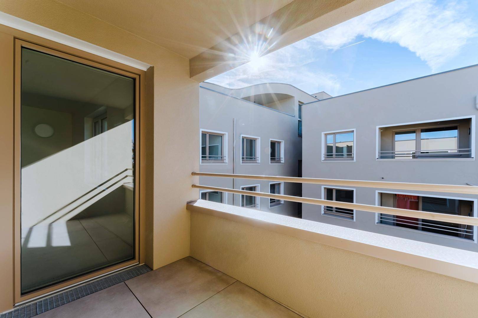 Sonniger Balkon mit Fliesenboden und Blick in den Innenhof des Gebäudes.