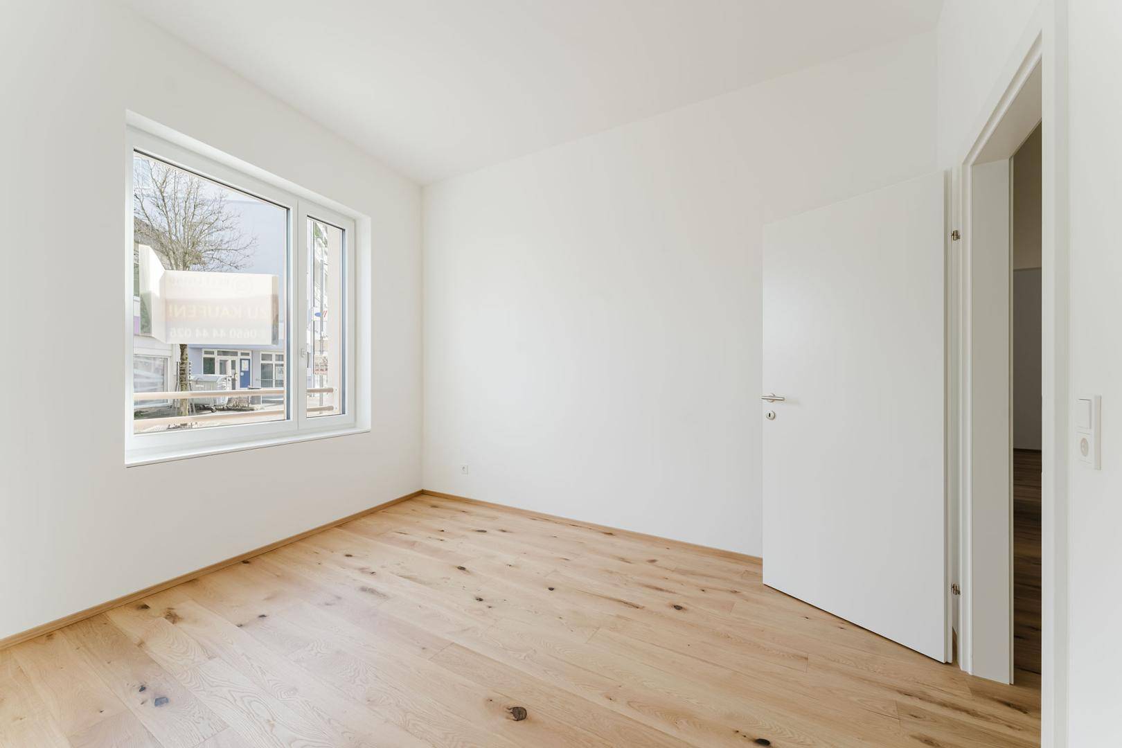 Helles, leeres Zimmer mit Holzboden und großem Fenster, ideal als Schlaf- oder Arbeitszimmer.