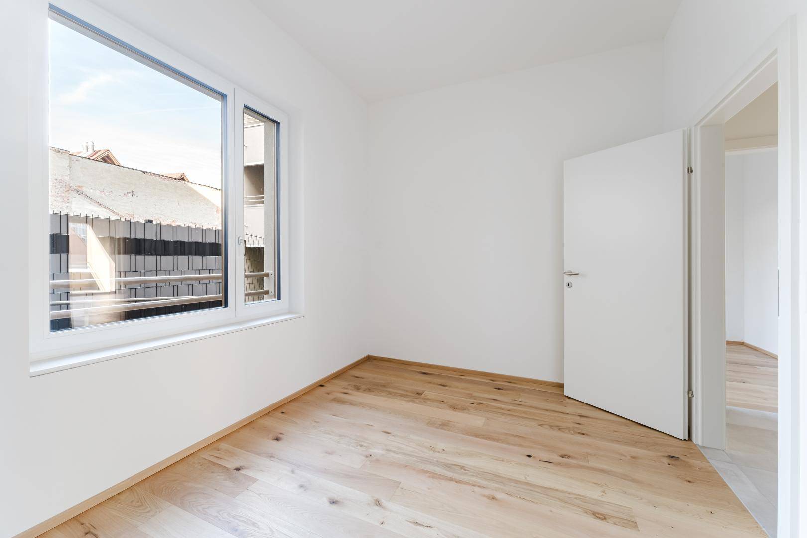 Leeres Zimmer mit hellem Holzboden und großem Fenster, ideal als Schlaf- oder Arbeitszimmer nutzbar.