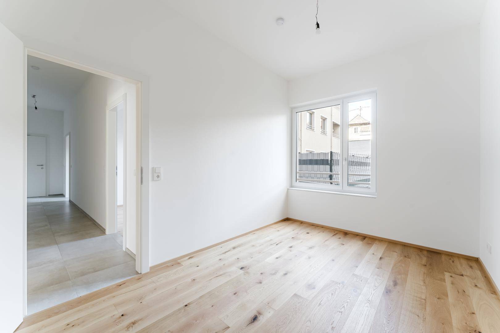 Helles Zimmer mit Holzboden und Fenster, das zum gefliesten Flur führt, in neuwertigem Zustand.