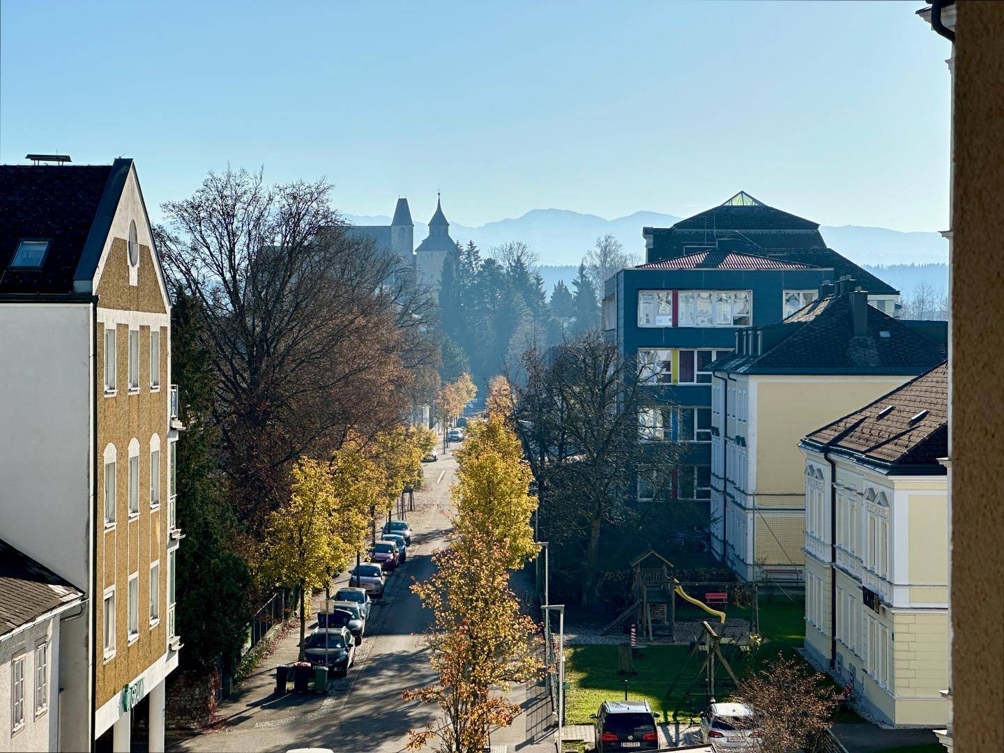Herbstlicher Ausblick auf eine belebte Straße mit bunten Bäumen und einer Bergkette im Hintergrund.