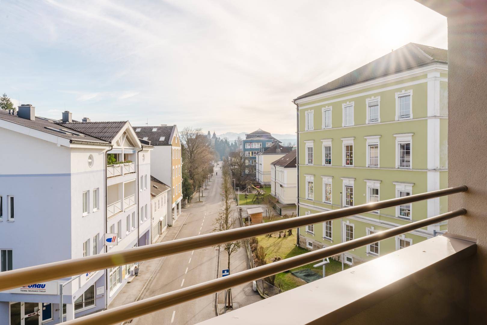 Blick vom Balkon auf eine ruhige Straße mit Gebäuden und die umliegende Berglandschaft unter strahlendem Himmel.