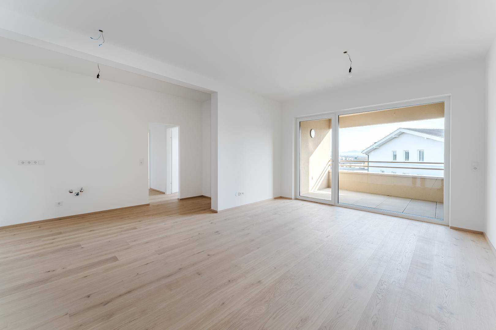 Helles, leeres Wohnzimmer mit hellem Holzfußboden und großem Schiebetürzugang zu einem Balkon oder einer Terrasse.