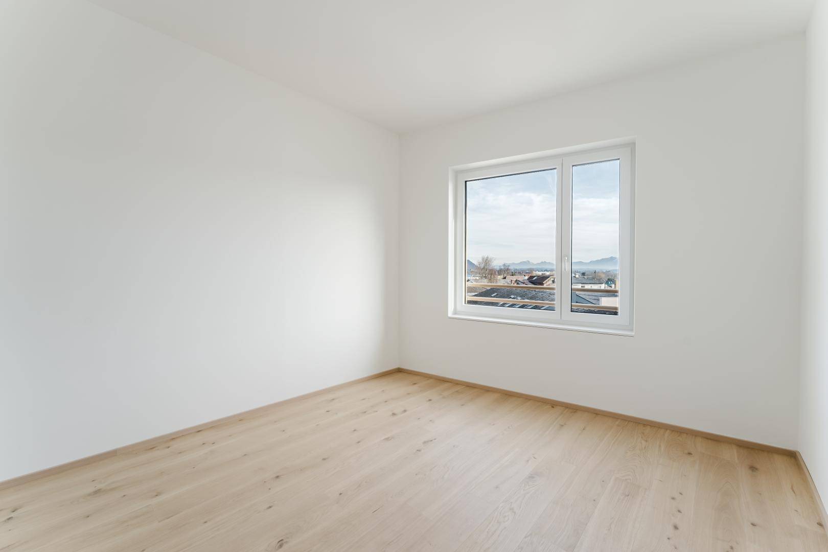 Helles, leeres Zimmer mit hellem Holzfußboden und einem Fenster mit Blick nach draußen.
