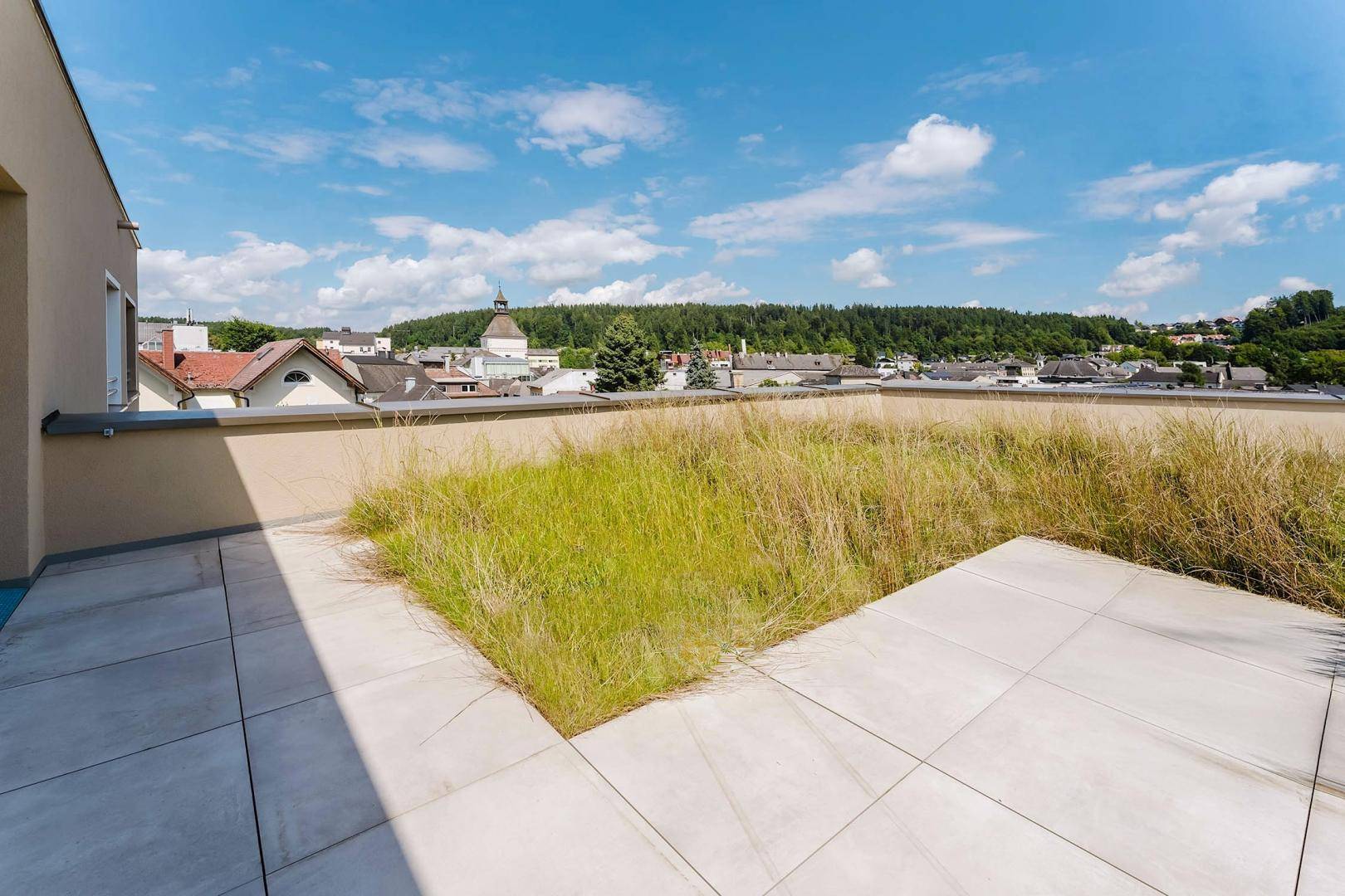 Teilweise begrünte Dachterrasse mit Fliesenboden und weitem Blick über die Stadt.